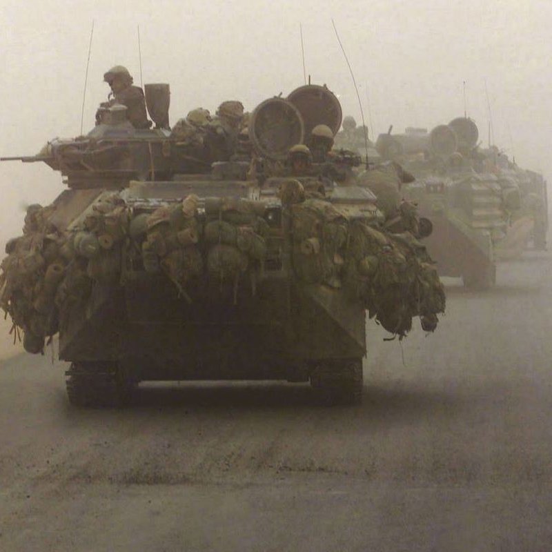 La imagen muestra un convoy militar en un ambiente polvoriento y desértico. Se pueden ver varios vehículos blindados, posiblemente tanques, avanzando en formación. Los vehículos están cargados con equipos y suministros, lo que sugiere que están en una misión de combate o de suministro. La visibilidad es limitada debido a la niebla o polvo, lo que añade una atmósfera tensa a la escena.