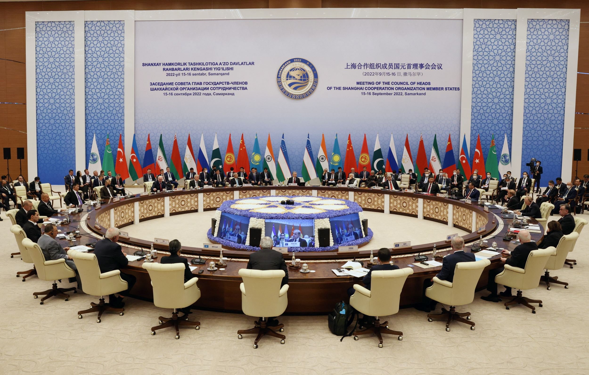 The image depicts a large conference room set up for a high-level meeting, with a circular table at the center surrounded by delegates. Various flags representing different countries are displayed around the room, indicating a multinational gathering. Attendees, likely officials or representatives, are seated and engaged in discussion. The backdrop features stylized graphics and text, suggesting it is related to a significant international conference, possibly focusing on cooperation among member states. The atmosphere appears formal and collaborative.