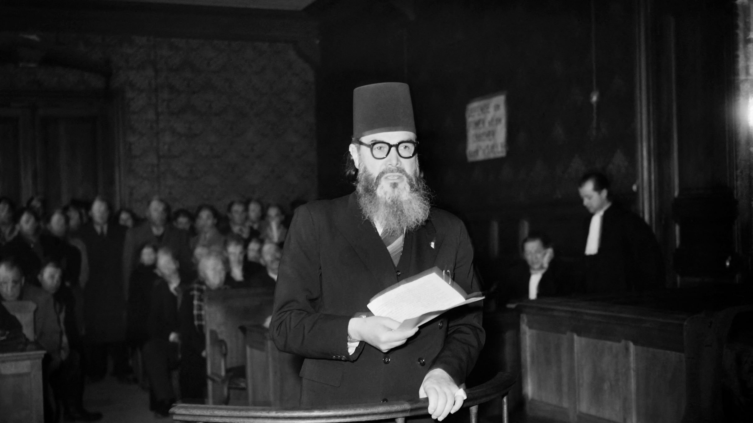 L'image montre un homme portant un turban noir et des lunettes, se tenant debout dans une salle d'audience. Il lit un document, tandis qu'un groupe de personnes l'écoute attentivement en arrière-plan. L'environnement est sombre, avec des boiseries et des bancs de juge visibles. Une personne en toge est également présente, ajoutant à l'atmosphère judiciaire de la scène.