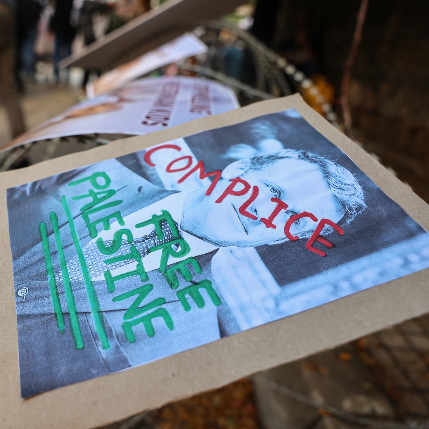 در تصویر، تعدادی از پوسترها روی یک نرده یا حصار قرار گرفته‌اند. یکی از این پوسترها تصویری از یک فرد همراه با نوشته‌هایی به زبان انگلیسی را نشان می‌دهد. نوشته‌های روی پوستر شامل کلماتی چون "COMPICILE" و "FREE PALESTINE" است. طراحی این پوستر به طور واضح به تضادهایی که بر سر مسائل فلسطین وجود دارد، اشاره دارد. زمینه‌ای غیررسمی و معترضانه به نظر می‌رسد و نشانه‌های حمایت از فلسطین را به تصویر می‌کشد.