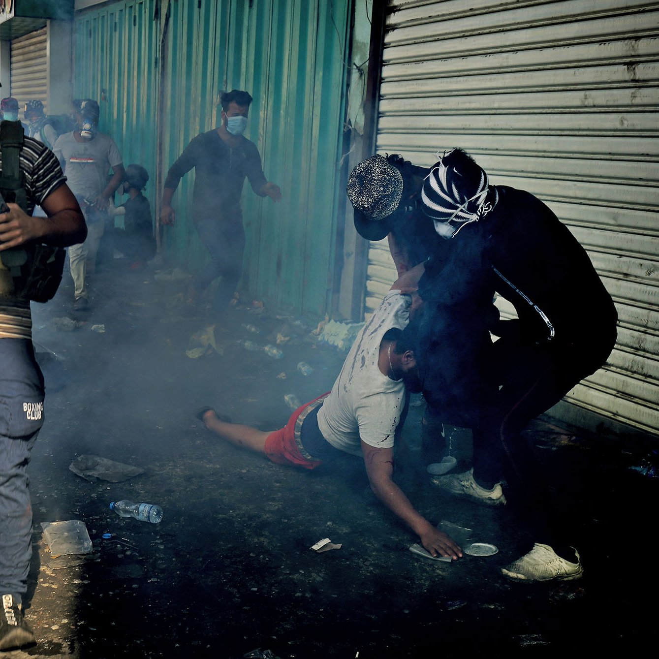 تصویر نشان‌دهنده یک صحنه پرتنش و شلوغ است. در foreground، یک مرد در حال دویدن به سمت دوربین است و ظاهراً در حال فرار از یک موقعیت خطرناک است. او کلاه‌خود و جلیقه نظامی به تن دارد. در پس‌زمینه، چند نفر دیگر دیده می‌شوند که در حال تعامل با یکدیگرند، یکی از آنها به زمین افتاده است. اطراف این افراد، دود و بخار به‌دلیل درگیری یا آشوب وجود دارد و زمین مملو از زباله و اشیای مختلف است. صحنه به وضوح فضای اضطراب و تنش را منتقل می‌کند.