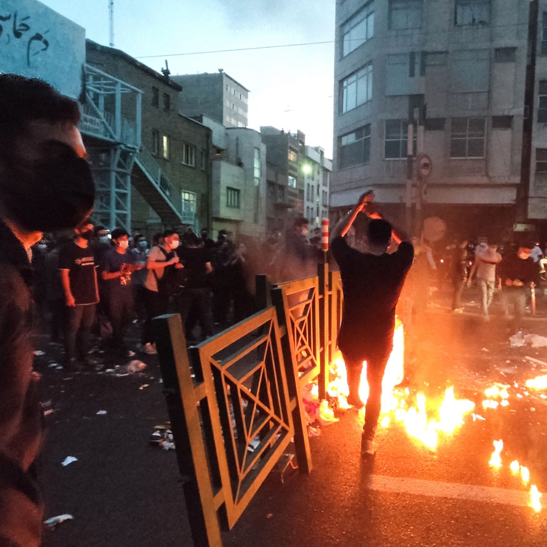 L'image montre une scène dynamique dans une rue urbaine, probablement lors d'une manifestation. On peut voir plusieurs personnes, dont l'une est au premier plan, se tenant près de flammes qui brûlent sur la route. L'atmosphère semble tendue, avec des barrières et des débris éparpillés. La lumière des flammes éclaire les visages des manifestants, qui portent des masques. En arrière-plan, on aperçoit des bâtiments urbains et une lumière tamisée, ce qui donne une impression d'urgence et de mouvement.