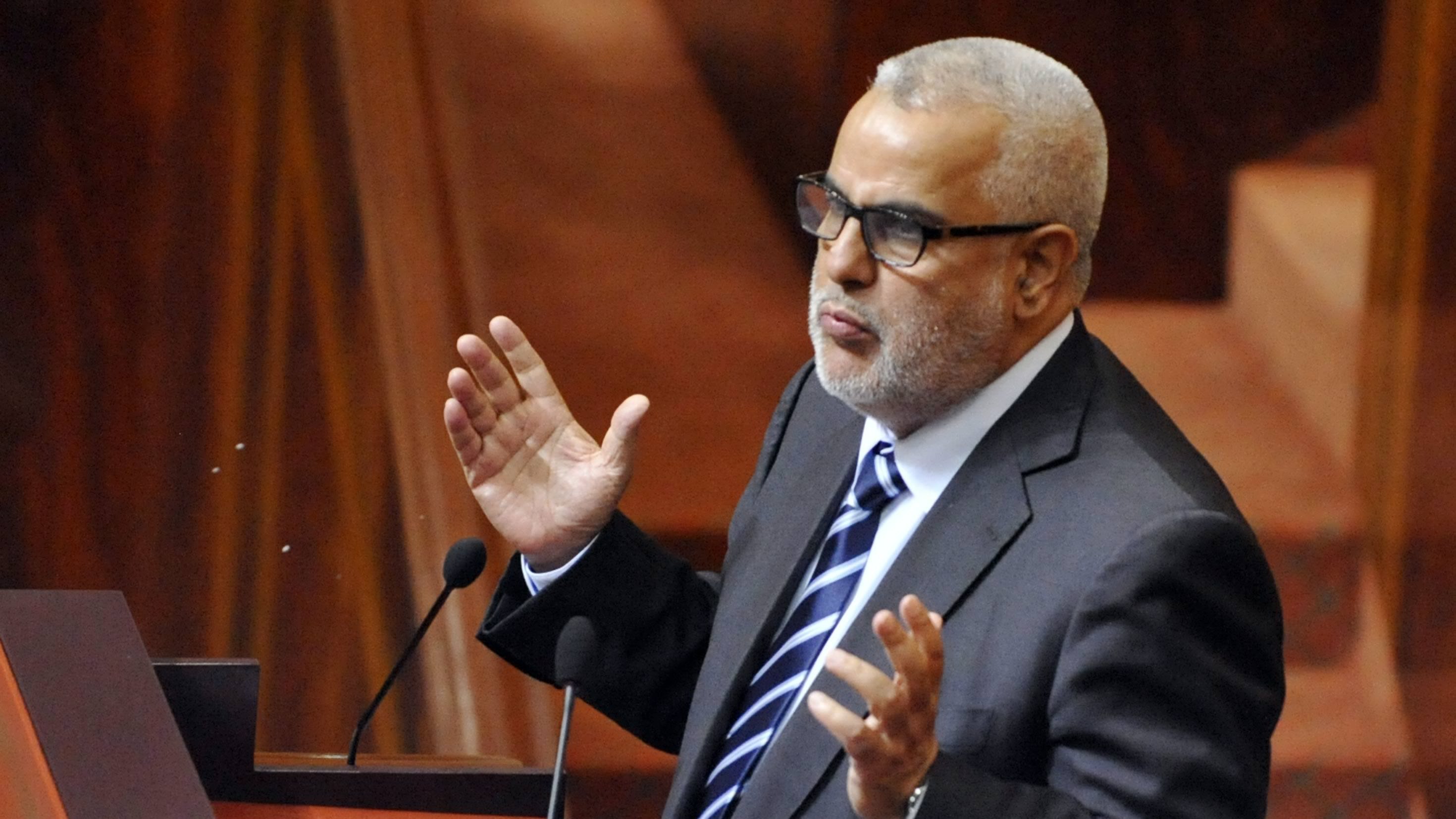 L'image montre un homme debout devant un pupitre, probablement en train de s'exprimer lors d'une réunion ou d'une séance législative. Il porte un costume sombre avec une chemise et une cravate à rayures. Son attitude et ses gestes suggèrent qu'il est engagé dans un discours ou une argumentation. L'environnement semble être celui d'une assemblée, avec des éléments de décor en bois et des sièges visibles en arrière-plan.