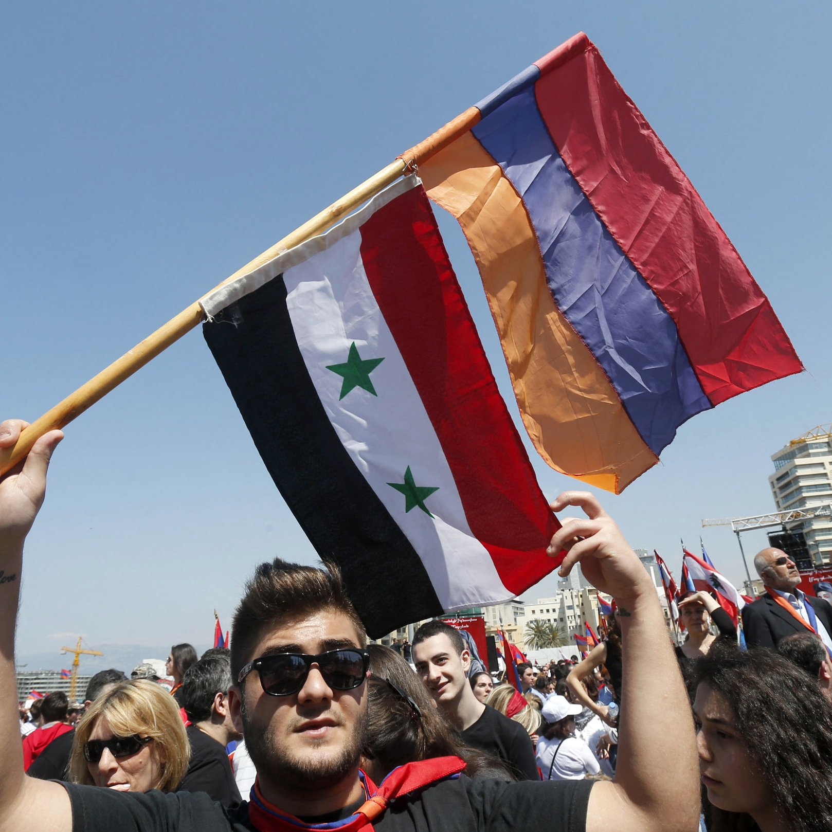 تظهر الصورة شخصًا يحمل عدة أعلام، بما في ذلك علم سوريا وعلم أرمينيا، في مكان يتجمهر فيه عدد من الناس. يبدو أن الجو يعكس حماسًا وطنيًا، حيث يرتدي العديد من المشاركين في المظاهرة ملابس تحمل ألوانًا أو رموزًا وطنية. الخلفية تحتوي على مباني حديثة، مما يشير إلى مكان حضري. السماء صافية، مما يضيف إلى الأجواء العامة.