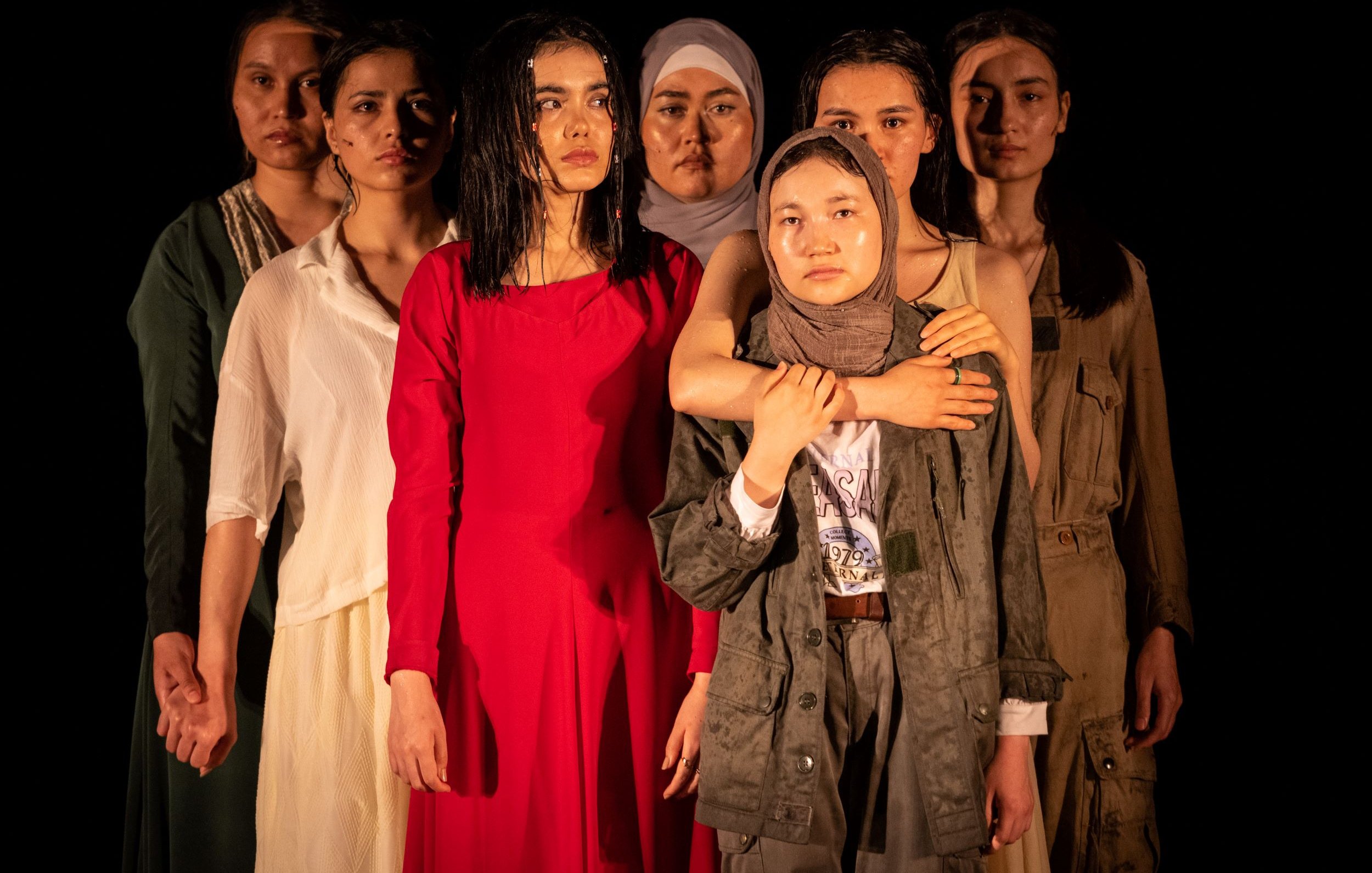 L'image montre un groupe de sept femmes, chacune portant des vêtements différents. Elles se tiennent en rang, avec des expressions sérieuses sur leurs visages. La femme au centre, vêtue d'une robe rouge, attire particulièrement l'attention. Les autres femmes, portant des vêtements variés allant de styles modernes à plus traditionnels, affichent une diversité de couleurs et de textures. L'éclairage met en valeur leurs visages et leurs postures, créant une atmosphère à la fois forte et émotive.