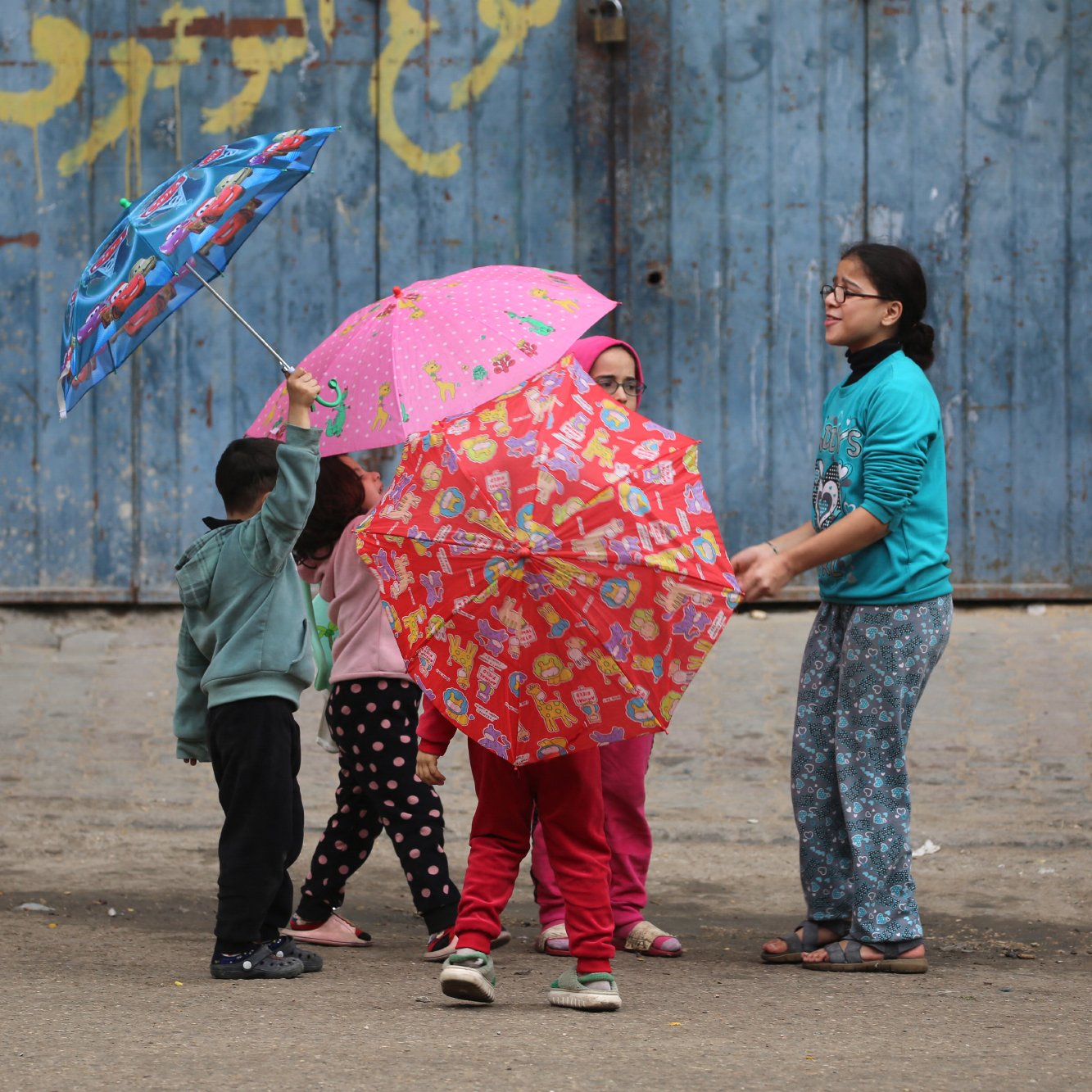 L'immagine mostra un gruppo di bambini che giocano insieme in strada, mentre tengono in mano degli ombrelli colorati. I bambini indossano abiti semplici e variopinti, e sembrano divertirsi sotto un muro blu con scritte. La scena trasmette gioia e convivialità.