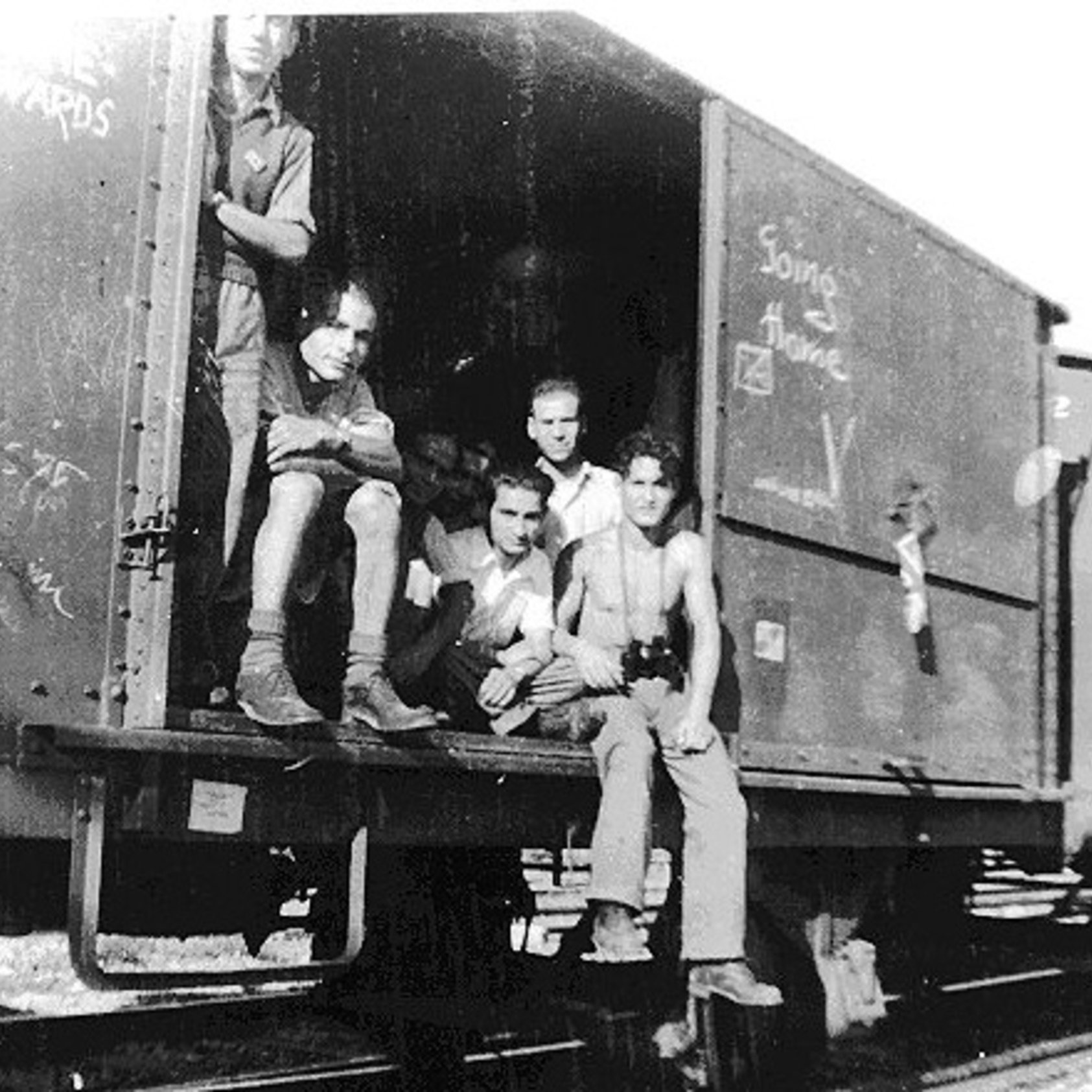 در این تصویر، گروهی از مردان جوان در یک واگن قطار نشسته‌اند. آنها در حالتی غیررسمی و دوستانه به نظر می‌رسند و برخی از آنها در حال استراحت هستند. دیوار واگن با نوشته‌ها و نقاشی‌هایی پوشیده شده است. تصویر به صورت سیاه و سفید گرفته شده و حس تاریخی و یادآور خاطرات گذشته را منتقل می‌کند. فضای تصویر نشان‌دهنده یک زمان و مکان خاص است که ممکن است در دوران جنگ یا شرایط خاصی باشد.