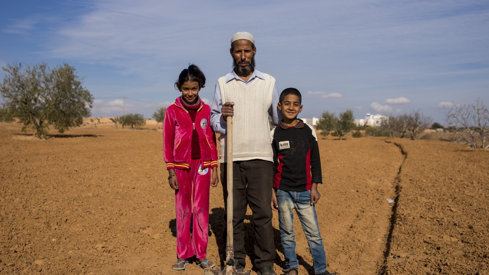 L'image montre un homme debout au milieu d'un paysage désertique, avec deux enfants près de lui, une fille et un garçon. L'homme tient une pelle et porte des vêtements simples. La fille est vêtue d'un ensemble rouge, tandis que le garçon porte un sweat-shirt noir avec des détails rouges. En arrière-plan, on peut voir quelques arbres éparpillés et un ciel bleu clair. Le sol est sec et nu, typique d'une région aride. L'ambiance semble conviviale, suggérant une journée de travail ou de récolte en famille.