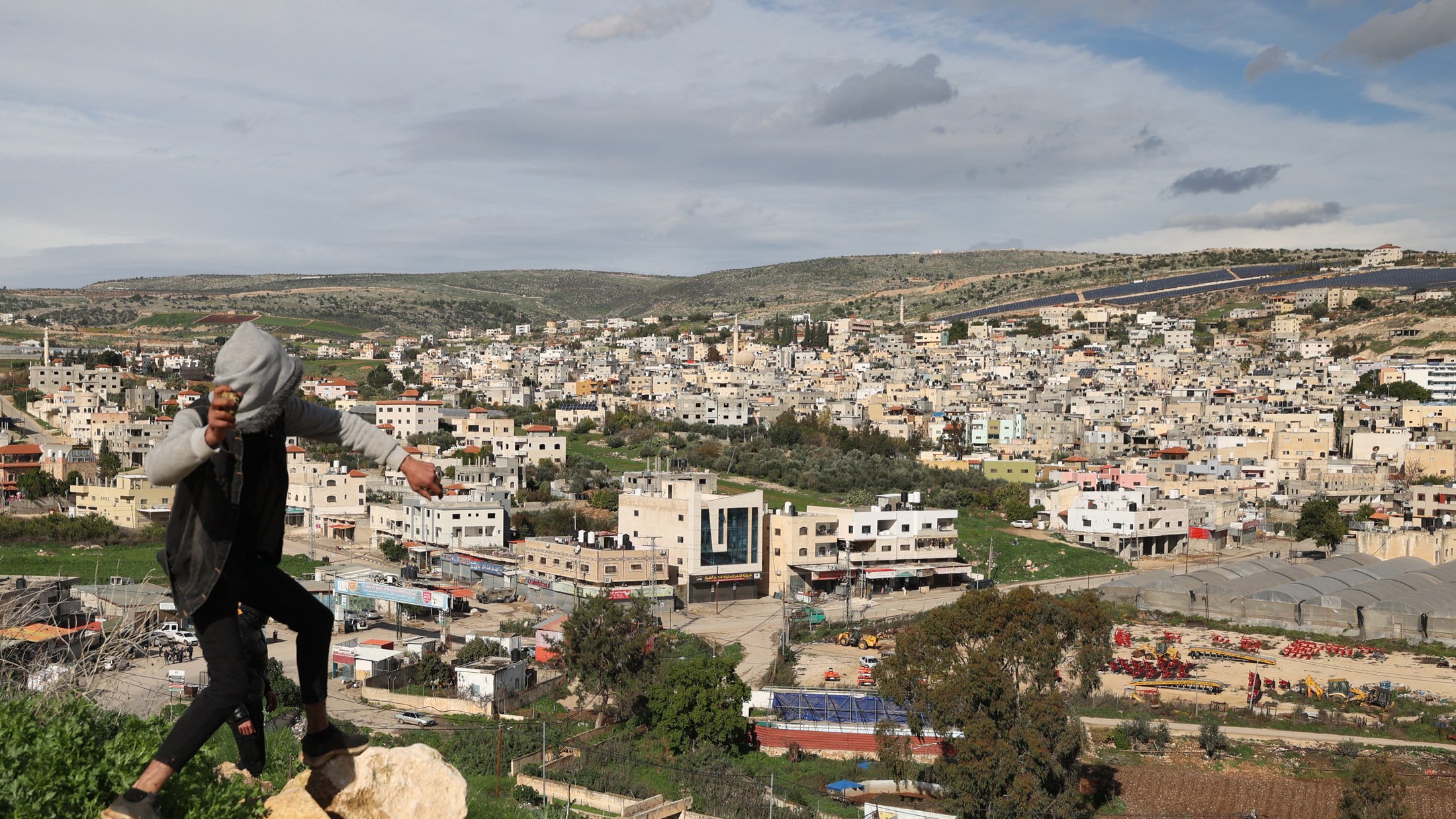 تظهر الصورة مشهداً طبيعياً لمدينة أو قرية، حيث تطل المباني السكنية على مساحات خضراء. في مقدمة الصورة، يوجد شخص يقف على صخرة، يبدو أنه في حالة حركية، بينما تظهر خلفه منظر عام للمدينة والمناطق المحيطة بها. السماء مليئة بالغيوم، مما يعكس أجواء متغيرة. تبرز في الصورة شوارع ومزارع، مما يعطي انطباعاً عن البيئة المحلية.