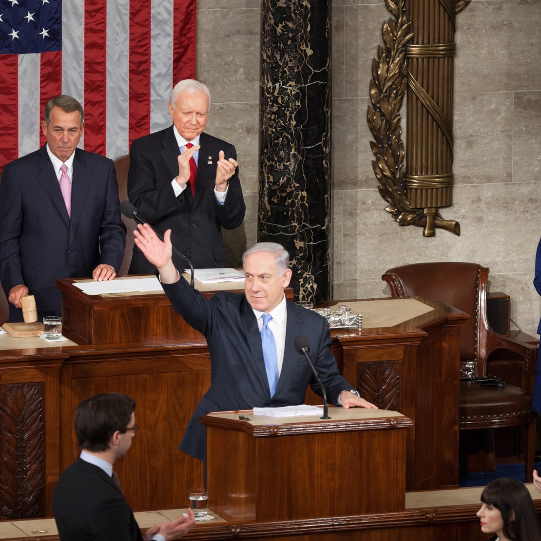 Cette image montre une scène dans une salle législative. Un homme se tient au pupitre, levant la main en signe de salut ou d'allocution. Derrière lui, plusieurs personnes, dont un homme plus âgé et une femme, l'écoutent attentivement. On peut également voir un drapeau américain en arrière-plan, ainsi que le mobilier typique d'une assemblée. L'atmosphère semble formelle et solennelle, suggérant un moment important, potentiellement un discours ou une présentation officielle.