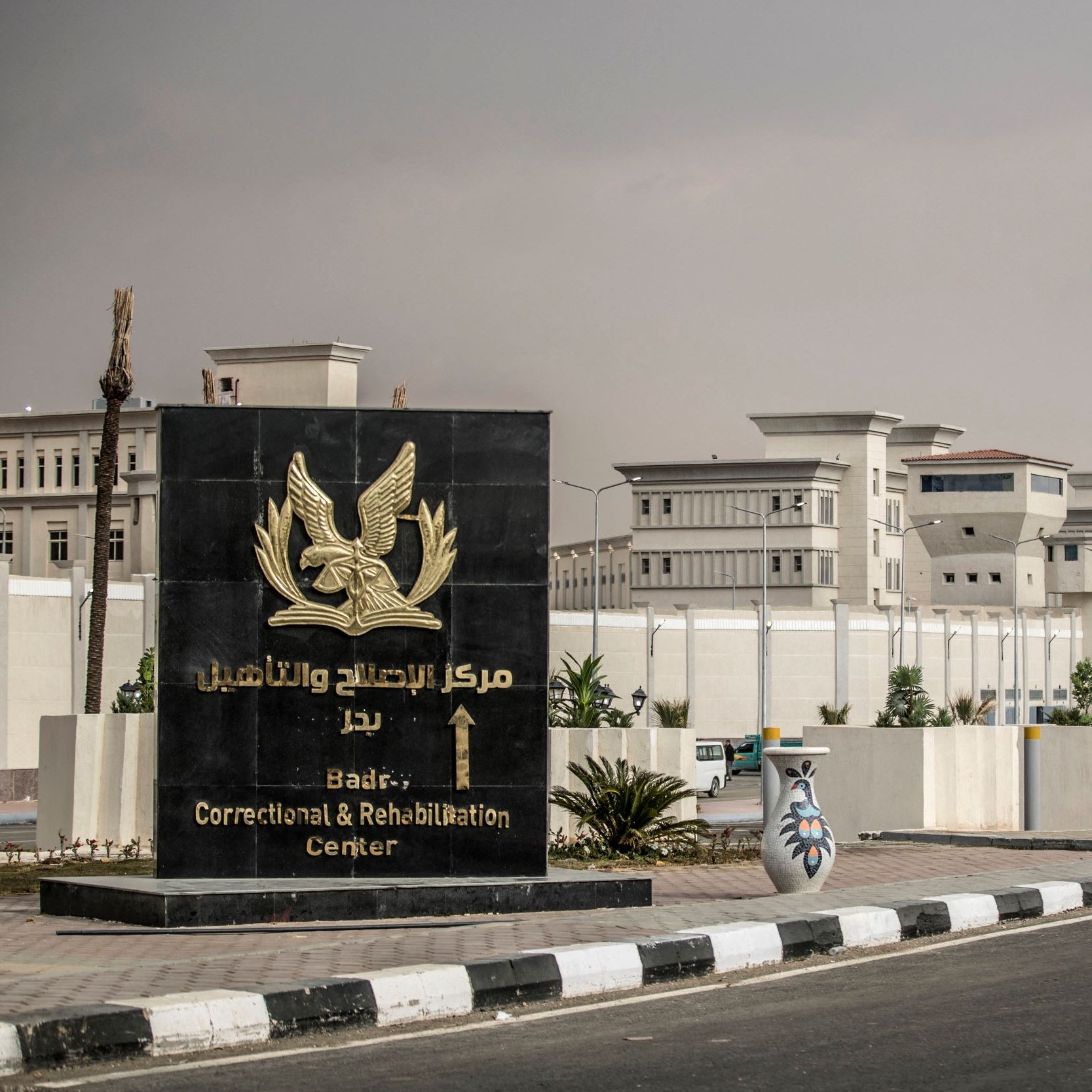 تظهر في الصورة لافتة كبيرة لمركز الإصلاح والتأهيل في بدر، حيث تحتوي على شعار أو رمز يرمز إلى المركز. المبنى خلف اللافتة يبدو حديثاً وعصرياً، محاطاً بسور أبيض ونباتات مصفوفة بشكل مرتب. الأجواء في الصورة تبدو هادئة، مع تواجد لبعض الأعمدة الضوئية على جانب الطريق.