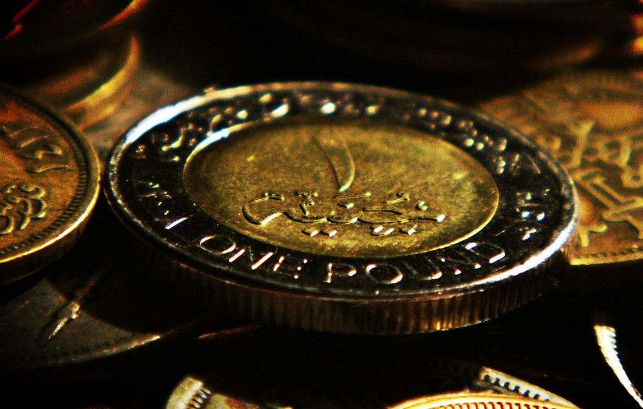 L'image montre plusieurs pièces de monnaie, avec une pièce de un pound en avant-plan. La pièce est en métal, et on peut voir des inscriptions en arabe sur sa surface, ainsi que les mots "ONE POUND". L'arrière-plan est flou, composé de plusieurs autres pièces de monnaie qui ajoutent à la richesse visuelle de la composition. Les pièces semblent avoir un éclat doré, ce qui attire l'attention sur le sujet principal.