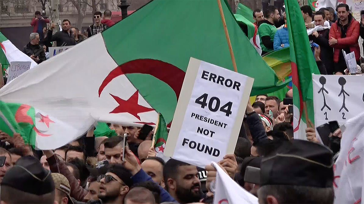 L'image montre une manifestation avec de nombreuses personnes brandissant des drapeaux algériens. Au premier plan, un manifestant tient une pancarte avec l'inscription "ERROR 404 PRESIDENT NOT FOUND", suggérant un mécontentement vis-à-vis de la situation politique. L'ambiance semble être celle d'une contestation, avec une foule réunie pour exprimer ses revendications.