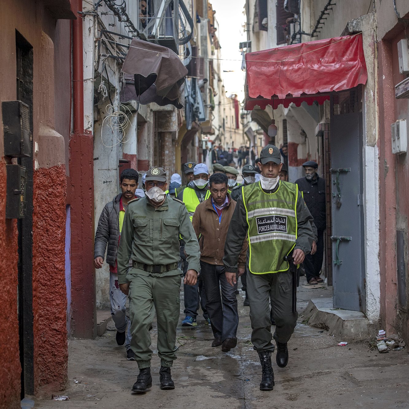 در این تصویر، یک کوچه باریک و قدیمی دیده می‌شود. در دو طرف کوچه، ساختمان‌ها با رنگ‌های مختلف نمایان هستند. گروهی از افرادی که لباس‌های فرم دارند، در حال قدم زدن در کوچه هستند. برخی از آن‌ها کلاه و ماسک به چهره دارند. محیط به نظر شلوغ و فعال می‌رسد و افراد مشغول به انجام کاری هستند. این تصویر فضایی محلی و اجتماعی را به نمایش می‌گذارد.