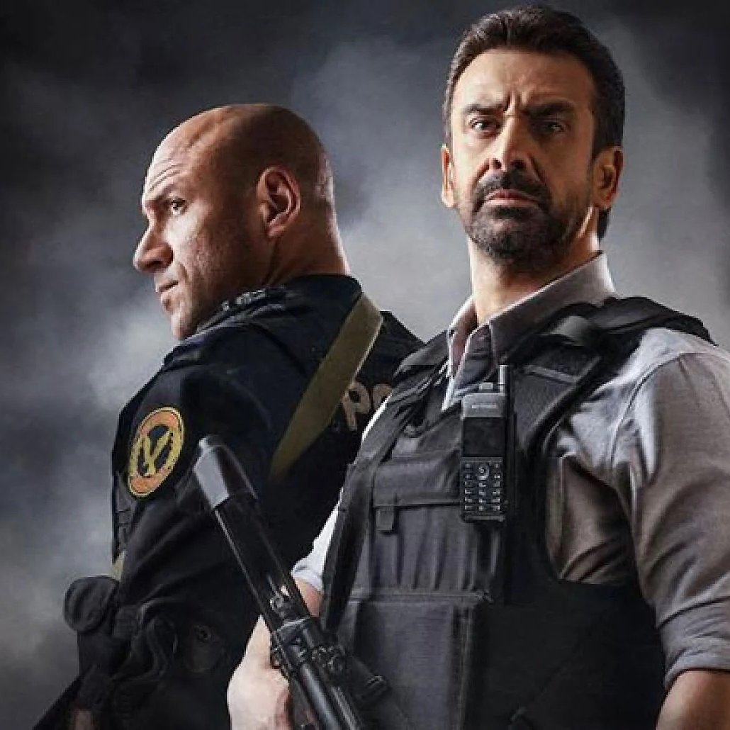 الصورة تظهر شخصين يرتديان زي الشرطة ويقفان معًا. أحدهما يبدو في وضعية أكثر تكاملًا، حيث يحمل سلاحًا ويعبر عن جدية معينة. الخلفية داكنة مما يعطي صورة درامية وينقل إحساسًا بالتوتر أو الإثارة. الشخصان يُظهران تعابير وجه تتسم بالتركيز، مما يوحي بأنهما في موقف يتطلب الحذر والانتباه.