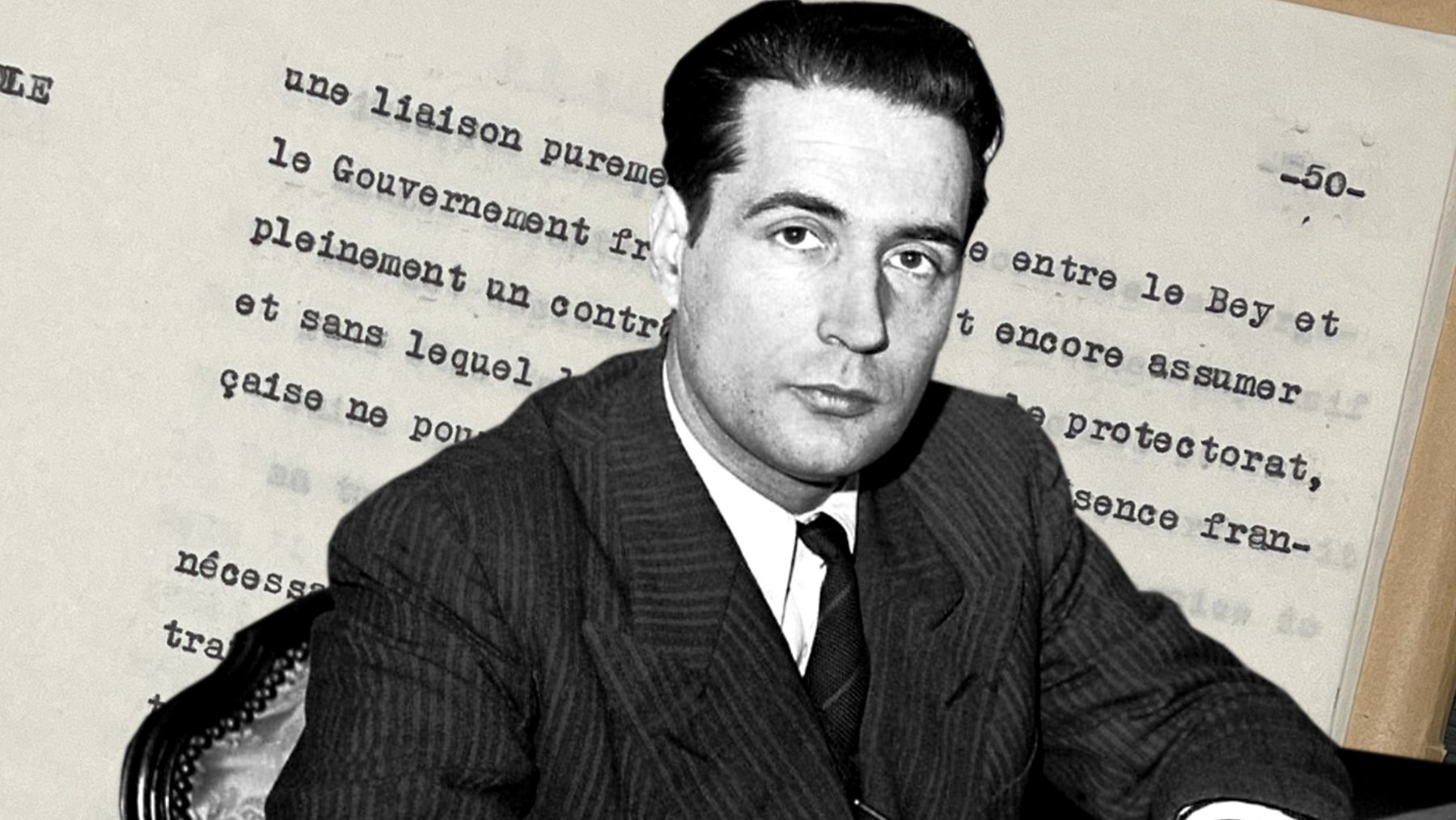 L'image montre un homme en costume assis à une table, tenant un stylo. En arrière-plan, on aperçoit des documents écrits, probablement des notes ou un texte officiel. L'homme a un regard sérieux et semble concentré sur son écrit. Son environnement suggère un contexte historique ou politique.