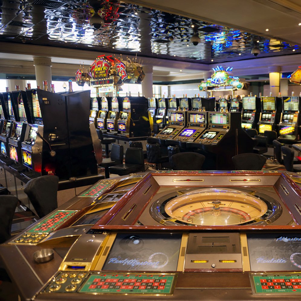 تصویر یک کازینو را نشان می‌دهد که در آن میزهای قمار و دستگاه‌های اسلات قرار دارند. در وسط تصویر، میز رولت با چرخ و دکمه‌های مختلف دیده می‌شود. در پس‌زمینه، دستگاه‌های اسلات و دیگر میزهای قمار به صورت منظم چیده شده‌اند. نورپردازی کازینو جذاب و رنگارنگ است و فضای عمومی، نوعی شلوغی و هیجان را به وجود آورده است.