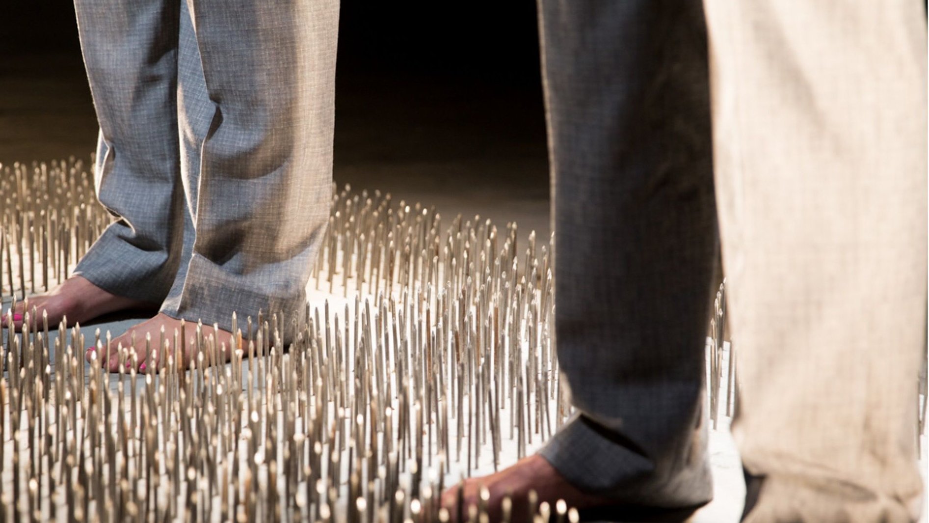 L'image montre deux personnes se tenant debout sur une surface couverte de nombreux picots métalliques. Les individus portent des pantalons clairs, et leurs pieds nus touchent les picots. La scène semble mettre en avant un contraste fort entre le confort des vêtements et l'inconfort potentiel de la surface sur laquelle ils se tiennent, créant une atmosphère à la fois intrigante et dérangeante.