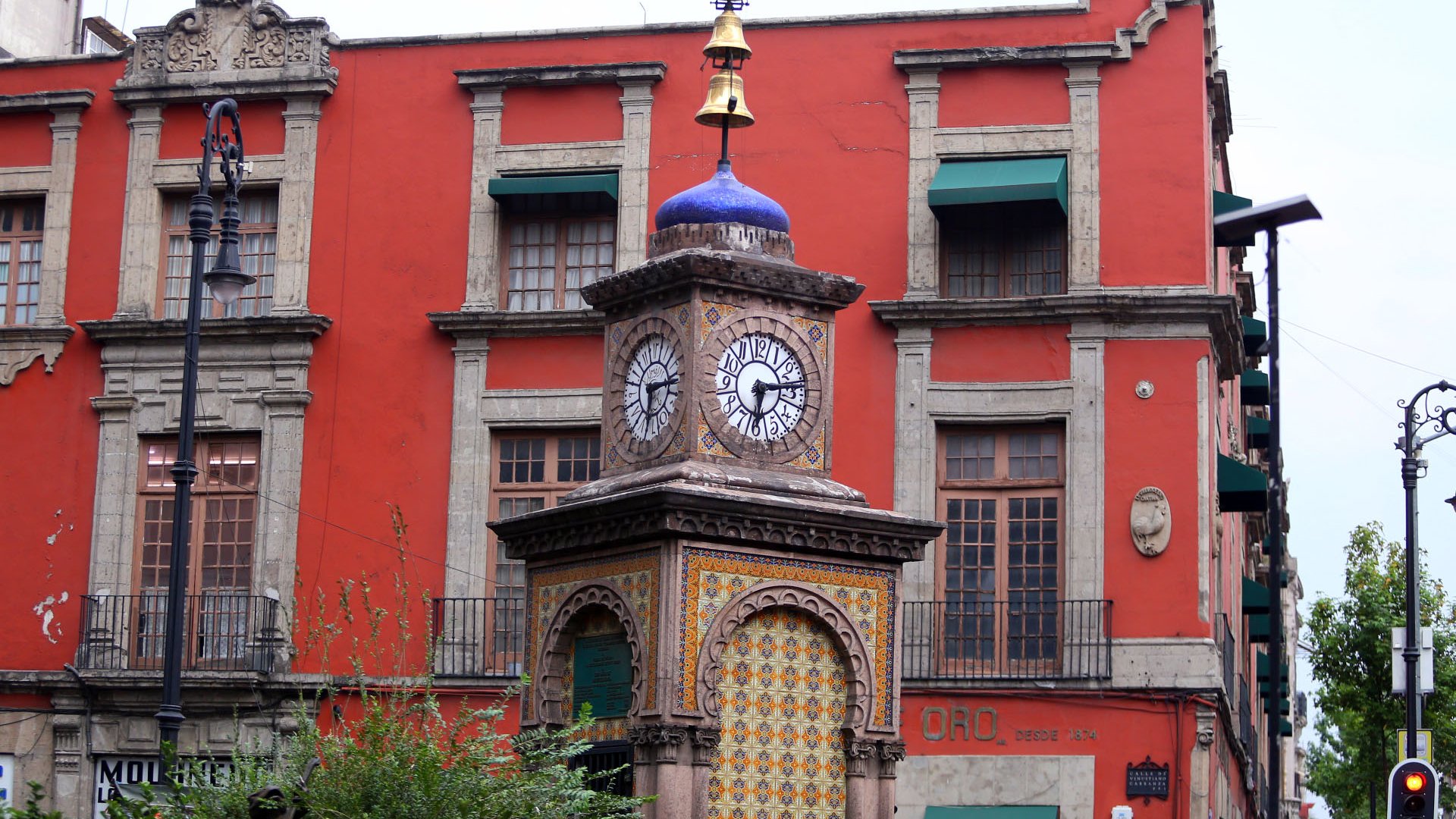 La imagen muestra un hermoso reloj de torre ubicado en una calle. La torre tiene una estructura ornamentada con un diseño colorido en su base y dos relojes visibles en los lados. Encima del reloj, hay una veleta dorada en la parte superior. Al fondo, se puede ver un edificio de color rojo con ventanas con contraventanas verdes. La arquitectura es de estilo colonial, con detalles decorativos en las fachadas. También hay una vegetación ligera en primer plano.