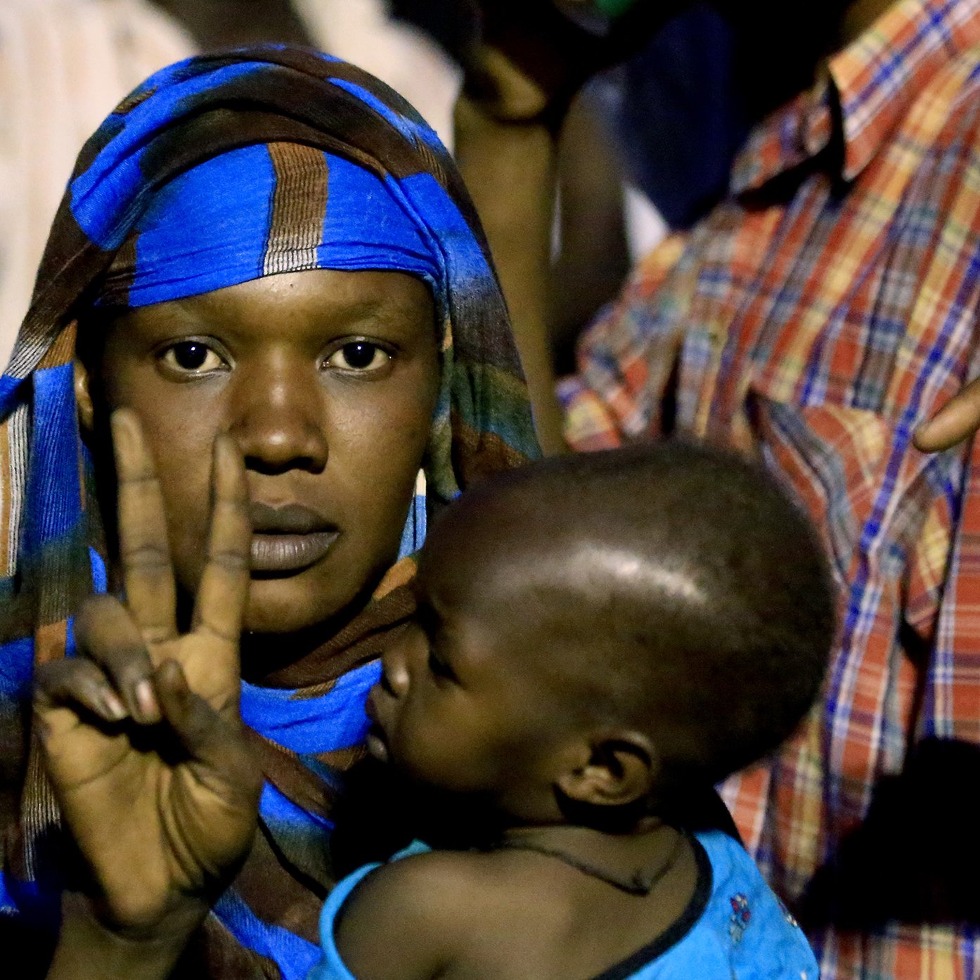 در این تصویر، زنی با یک شال رنگارنگ و نقوش آبی در حال نشان دادن علامت پیروزی (دو انگشت) است. او یک کودک را در آغوش دارد که به سمت او نگاه می‌کند. پس‌زمینه تصویر شامل افرادی است که در حال تماشا هستند و نور محیط کمی کم است. حالت زن نشان‌دهنده اعتماد به نفس و امیدواری است.