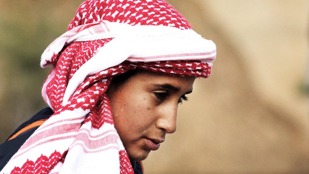L'image montre un jeune homme portant un keffieh, un traditionnel foulard arabe, qui est souvent orné de motifs rouges et blancs. Son expression semble pensive, et il a les cheveux bruns. L'arrière-plan est flou, indiquant un environnement naturel ou rural. Son regard est tourné légèrement vers le bas, ce qui ajoute une certaine introspection à son apparence.