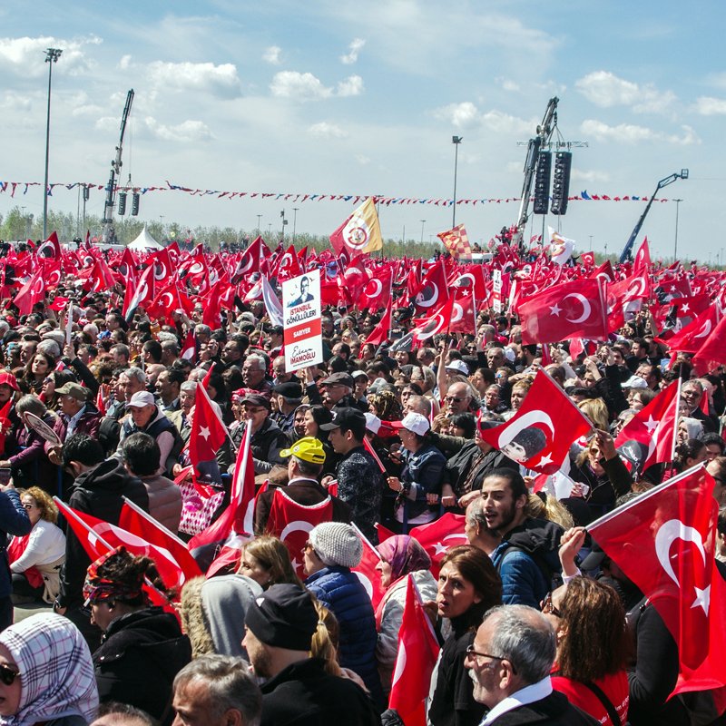The image shows a large crowd gathered in an outdoor setting, with many people holding Turkish flags. The atmosphere appears to be lively and energetic, with individuals visibly engaged in the event. Some are holding banners, while others are raising their flags high. The background features clear skies and some structures, possibly related to the event or gathering. Overall, it depicts a significant public demonstration or celebration, likely related to a national or cultural event in Turkey.