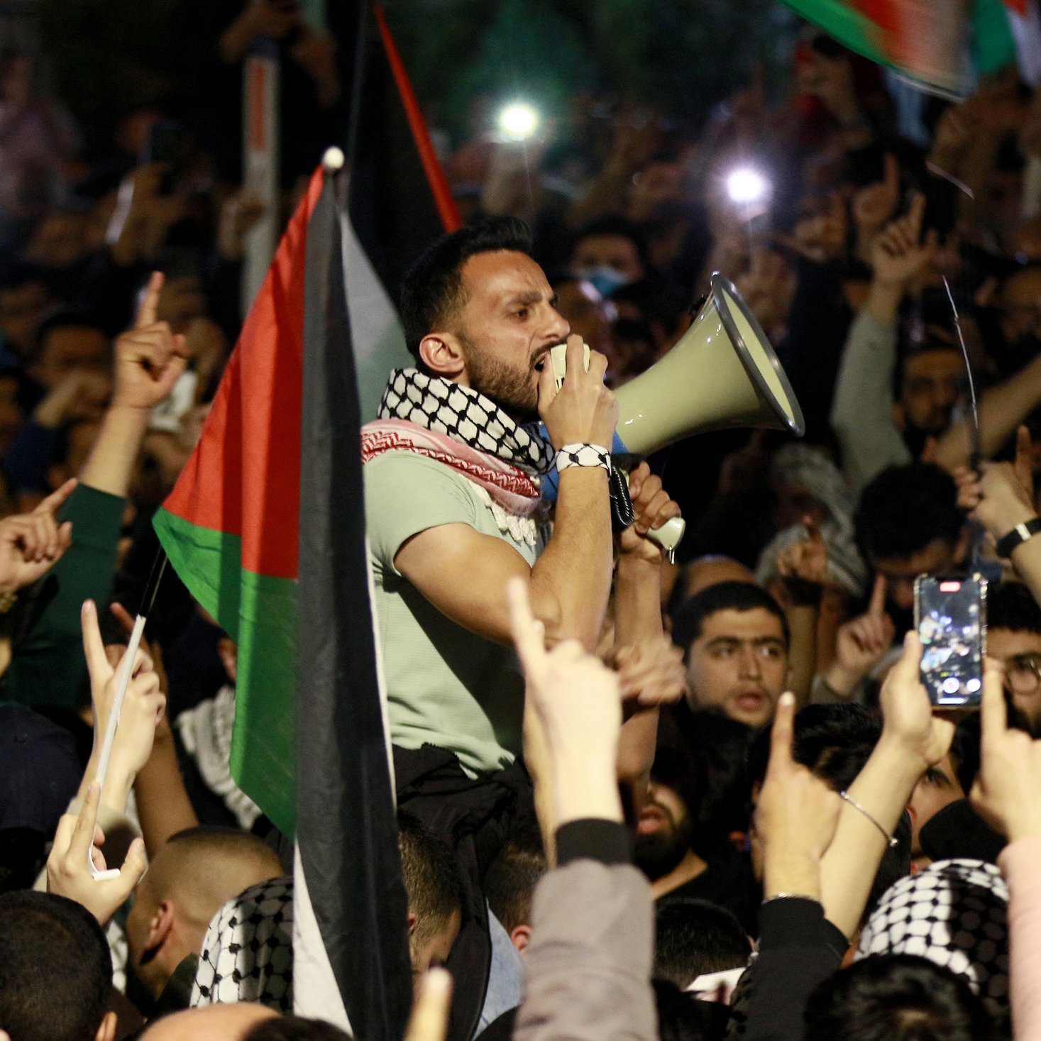 تصویر نشان‌دهنده جمعیتی از افراد است که در یک تجمع اعتراضی شرکت کرده‌اند. در مرکز تصویر، مردی با میکروفون در دست در حال سخنرانی یا فریاد زدن است. این فرد با پوششی که شامل شال معروف فلسطینی است، به نظر می‌رسد که به موضوعی مهم صحبت می‌کند. در پس‌زمینه، جمعیت زیادی با پرچم‌های فلسطین و دست‌هایی که به نشانه اعتراض یا حمایت بالا رفته‌اند، دیده می‌شود. این تصویر وضعیت هیجان‌انگیز و پرشور این تجمع را منتقل می‌کند.