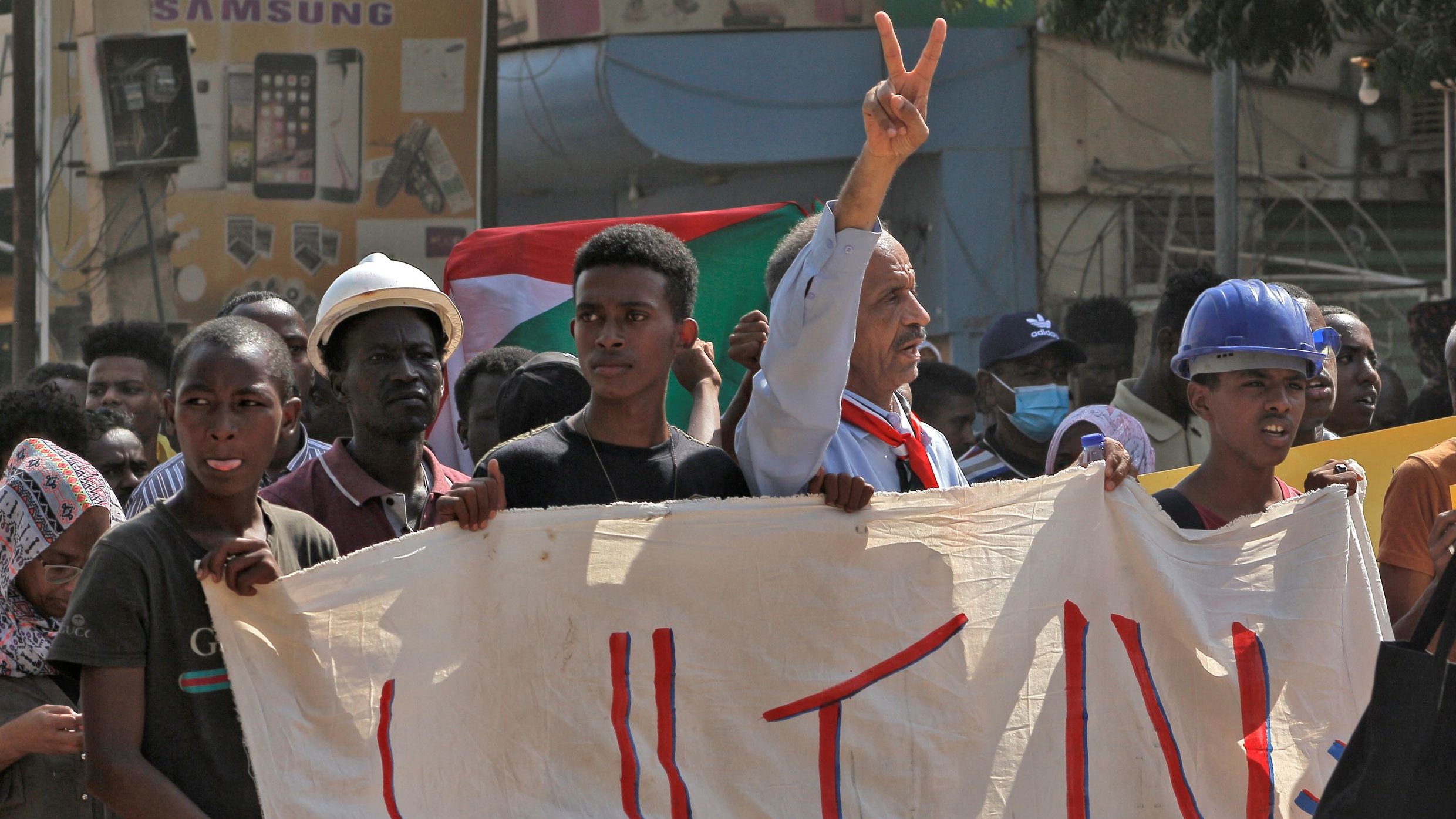 در این تصویر، گروهی از معترضان در حال برگزاری یک تظاهرات هستند. یکی از افراد، در حالی که علامت پیروزی را بالا برده، توجه را به خود جلب کرده است. دیگر شرکت‌کنندگان نیز با چهره‌های جدی و مصمم در کنار او ایستاده‌اند. در دست آنها یک بنر بزرگ دیده می‌شود که به زبان عربی نوشته شده و احتمالاً به اعتراضات مرتبط است. پس‌زمینه تصویر شامل ساختمان‌ها و تابلوهای تبلیغاتی است. احساس کلی تصویر نشان‌دهنده روحیه فعالان اجتماعی و خواسته‌های آنهاست.