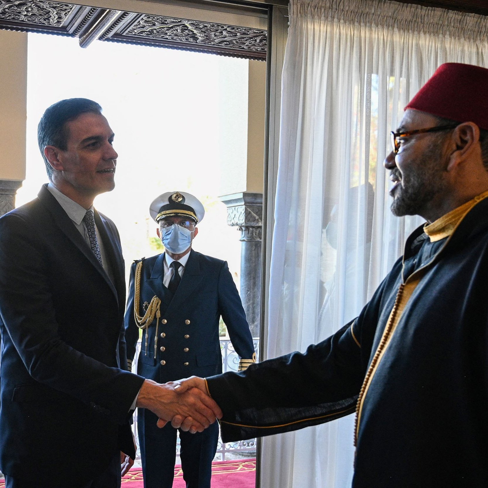 La imagen muestra a dos hombres dándose la mano en un entorno formal. Uno parece ser un líder o dignatario, vestido con un traje oscuro, mientras que el otro lleva una vestimenta tradicional, posiblemente un manto y un fez. En el fondo, se puede ver a un tercero, que es un guardia o oficial en uniforme, observando la escena. La decoración del lugar es elegante, con cortinas blancas y un entorno arquitectónico notable.