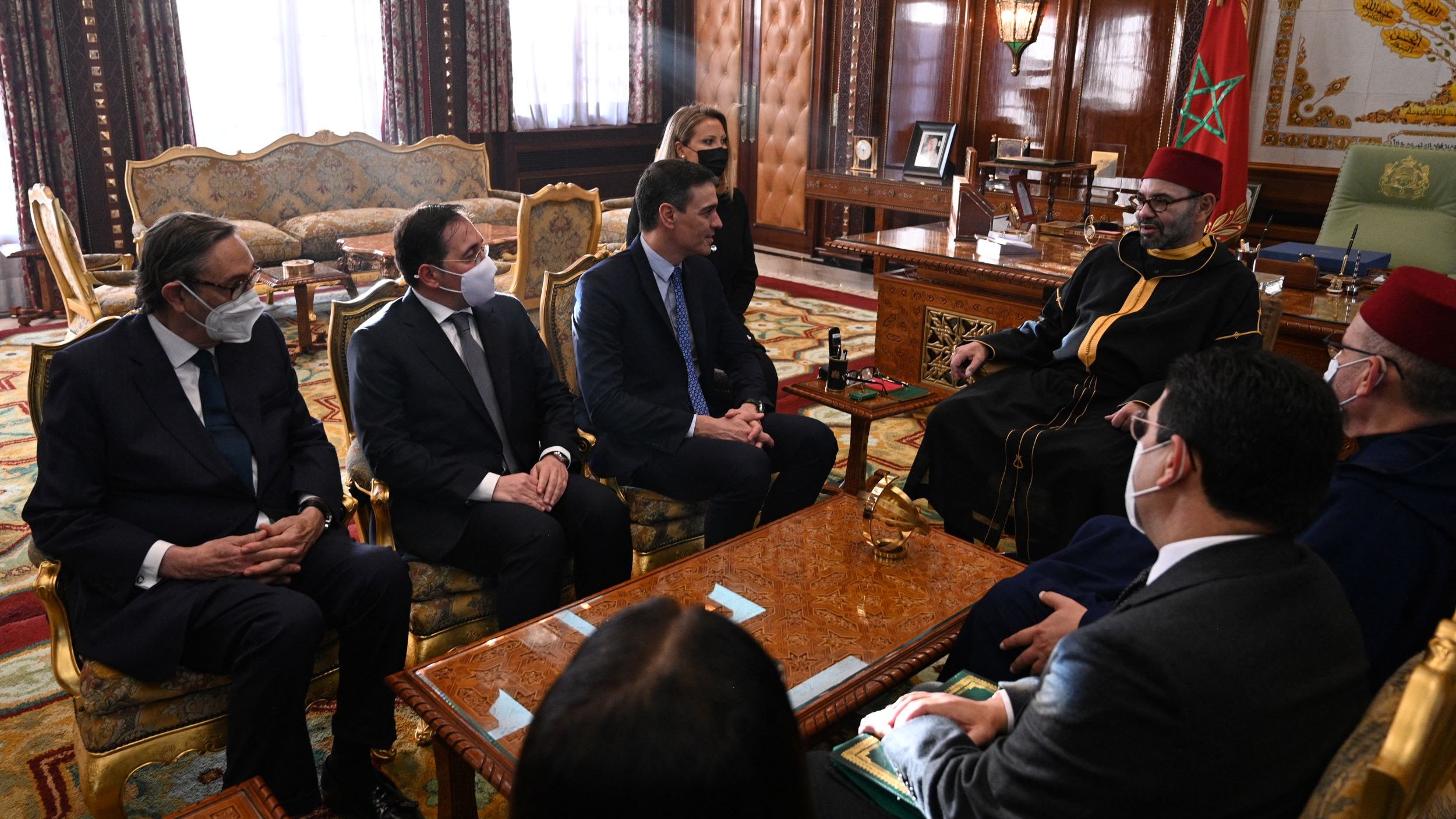 L'image montre une réunion dans un cadre officiel, probablement un bureau ou une salle de réunion importante. Plusieurs personnes en costumes sont assises autour d'une table, tandis qu'une autre personne, vêtue d'une tenue traditionnelle, est assise en face d'eux. Les participants portent des masques, ce qui indique que la réunion pourrait avoir lieu en période de pandémie. L'environnement est richement décoré, avec des éléments de design marocain et un mobilier en bois orné.