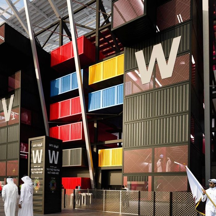 L'image montre une vue d'un bâtiment moderne, probablement un stade ou un centre événementiel, avec une architecture unique. Les murs extérieurs sont composés de containers colorés, alternant des teintes de jaune, rouge et bleu, donnant un aspect dynamique et contemporain. On distingue également des personnes en vêtements traditionnels se tenant près de l'entrée, ainsi que des éléments de sécurité. Le ciel est légèrement nuageux, ajoutant une ambiance particulière à la scène.