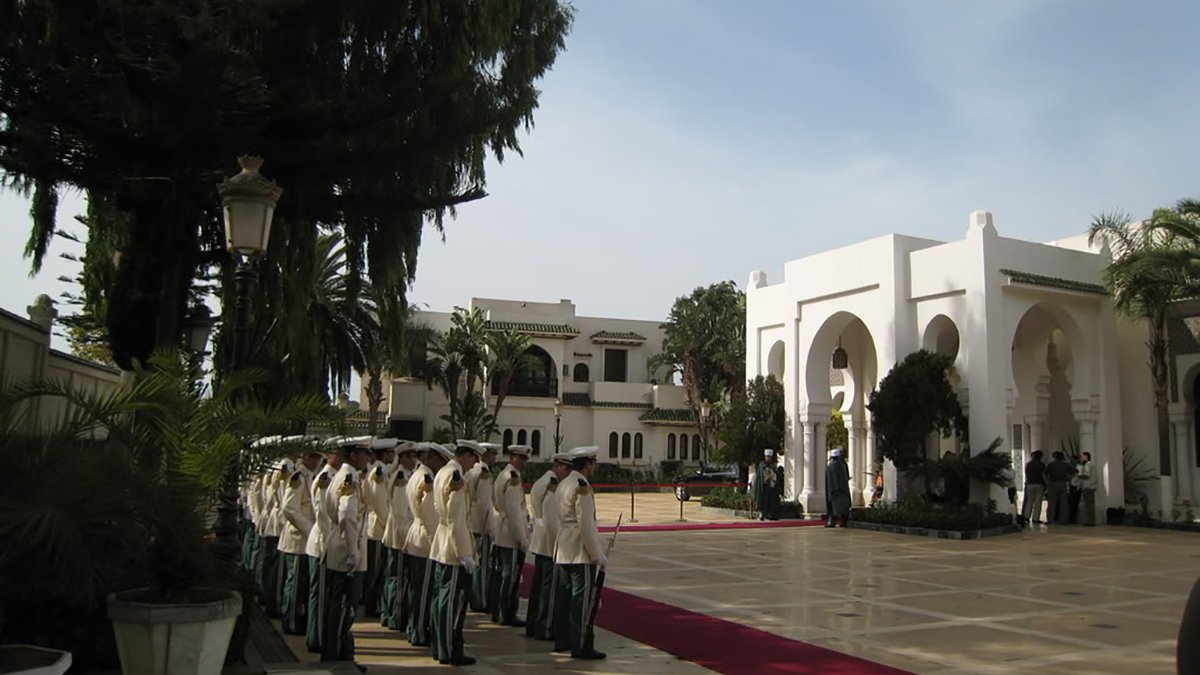 L'image montre une scène extérieure d'un bâtiment élégant, probablement un palais ou un hôtel de luxe. On peut voir une allée centrale avec un tapis rouge, entourée de verdure et de palmiers. À gauche, un groupe de personnes en uniformes formés, représentant une sorte de garde ou de cérémonie. En arrière-plan, on aperçoit un bâtiment majestueux avec des arches et des colonnes, suggérant une architecture raffinée. Le ciel est clair, ce qui ajoute à l'atmosphère solennelle de la scène.