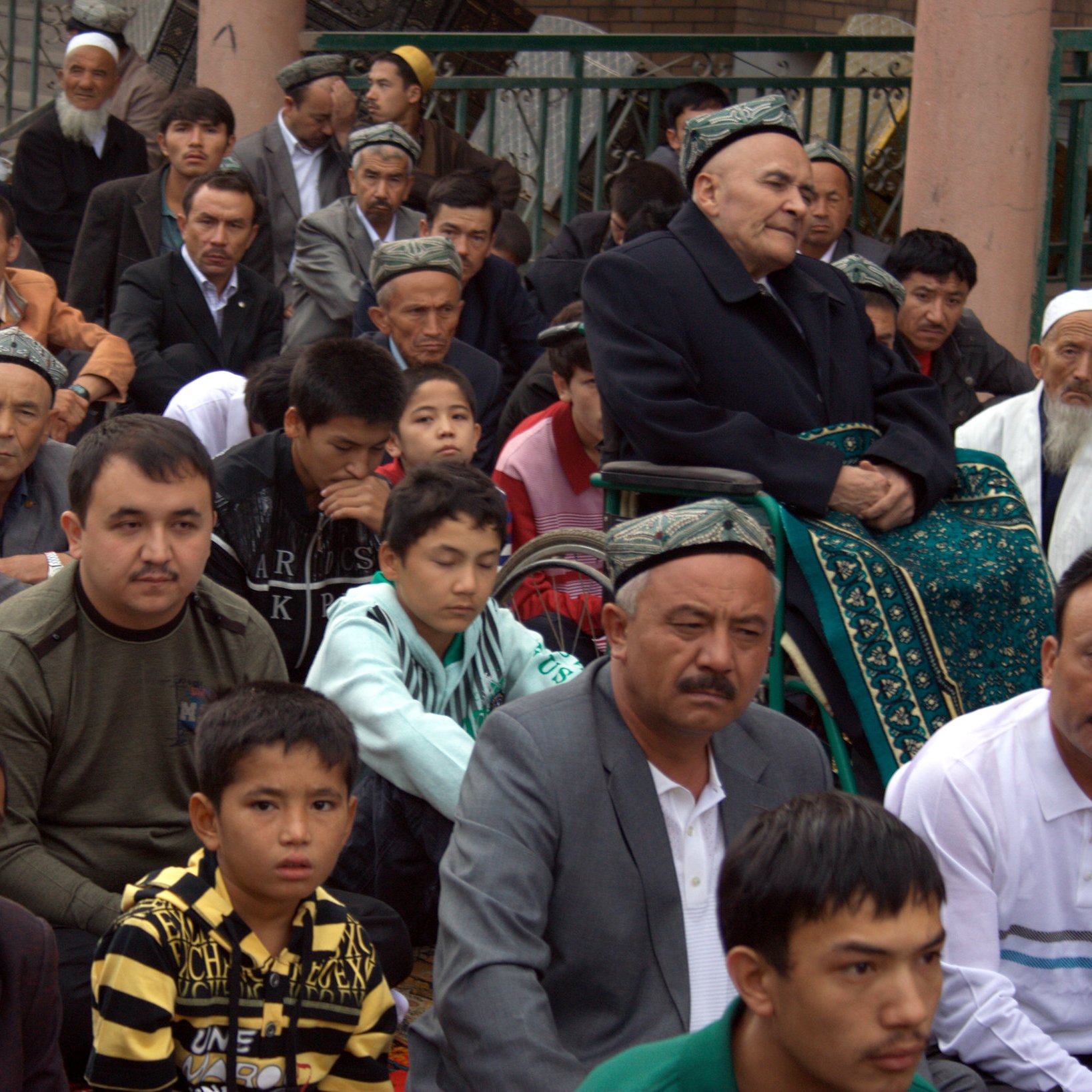 La imagen muestra a un grupo de personas sentadas en un ambiente al aire libre, posiblemente durante un evento comunitario o religioso. Se pueden ver hombres y niños, algunos de ellos vestidos con ropa tradicional. En el centro de la imagen, hay un hombre mayor sentado en una silla de ruedas, mirando hacia el frente. Los rostros muestran atención y respeto, reflejando la seriedad del momento. La escena parece estar en un lugar público, rodeado de otros espectadores.