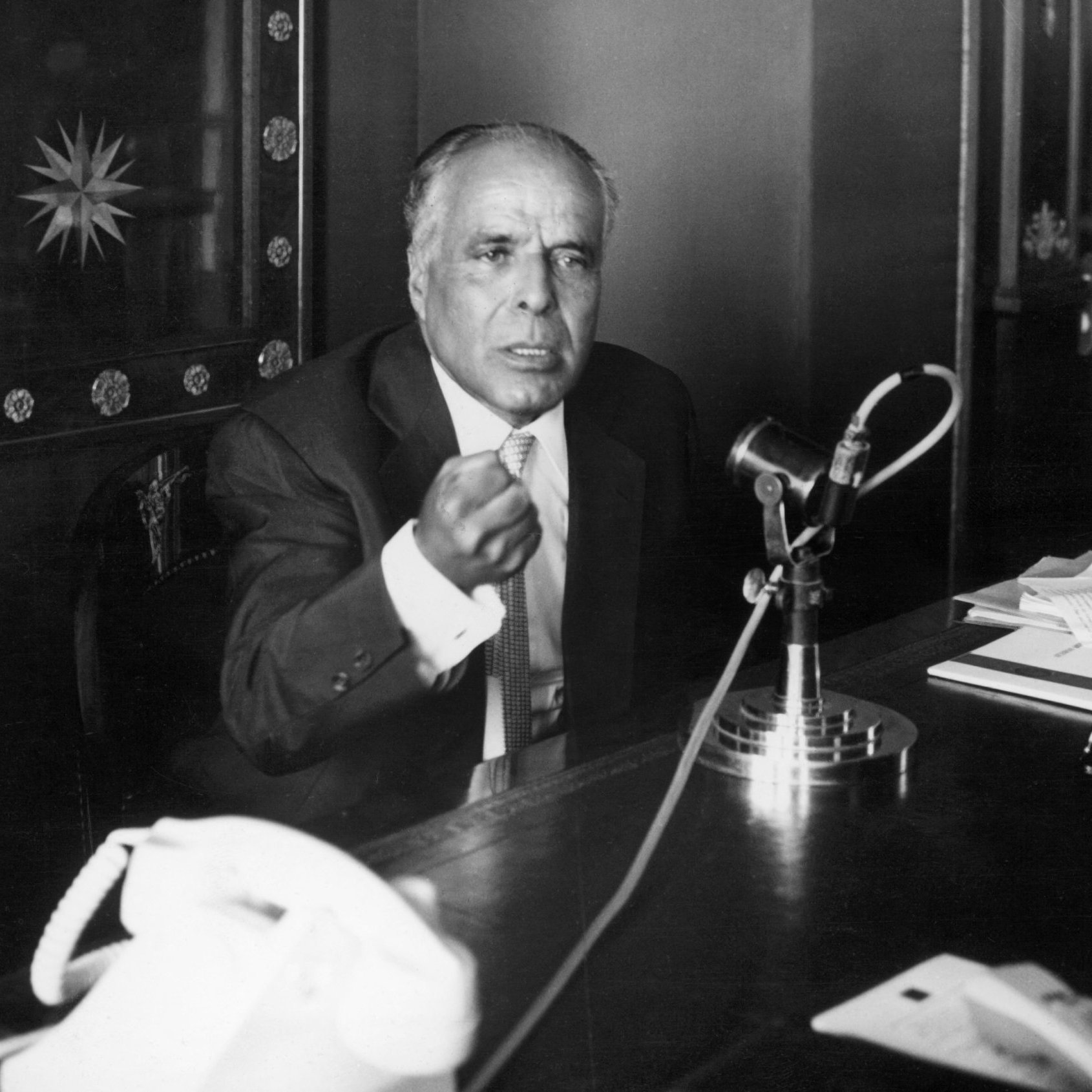 Homme en costume, gesticulant, devant un microphone et un bureau rempli de documents.