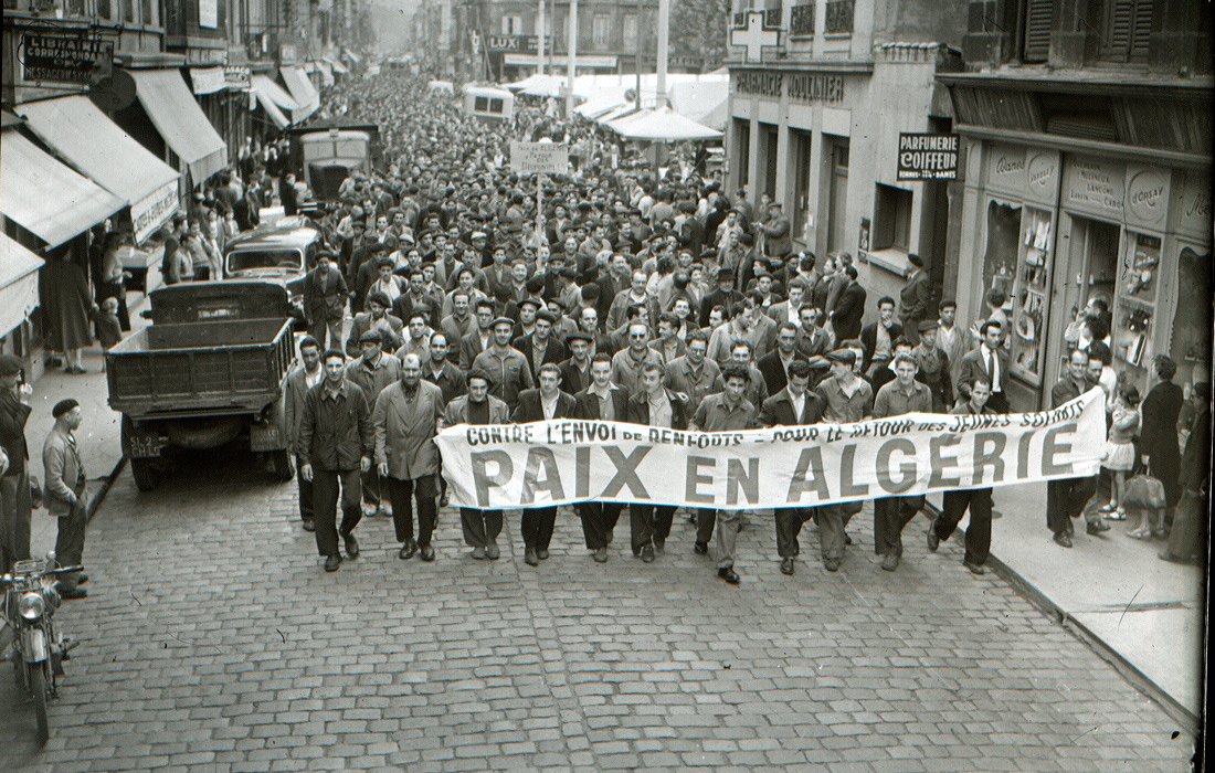 L'image montre une grande foule rassemblée dans une rue, probablement lors d'une manifestation. Les personnes marchent en portant une grande banderole qui indique "PAIX EN ALGÉRIE". On peut voir des hommes et quelques femmes, habillés de manière vestimentaire typique de l'époque. Des voitures et des commerces sont visibles en arrière-plan, ce qui donne une idée du contexte urbain. L'atmosphère semble sérieuse, suggérant que la manifestation est organisée autour d'un sujet important, en l'occurrence la paix en Algérie.