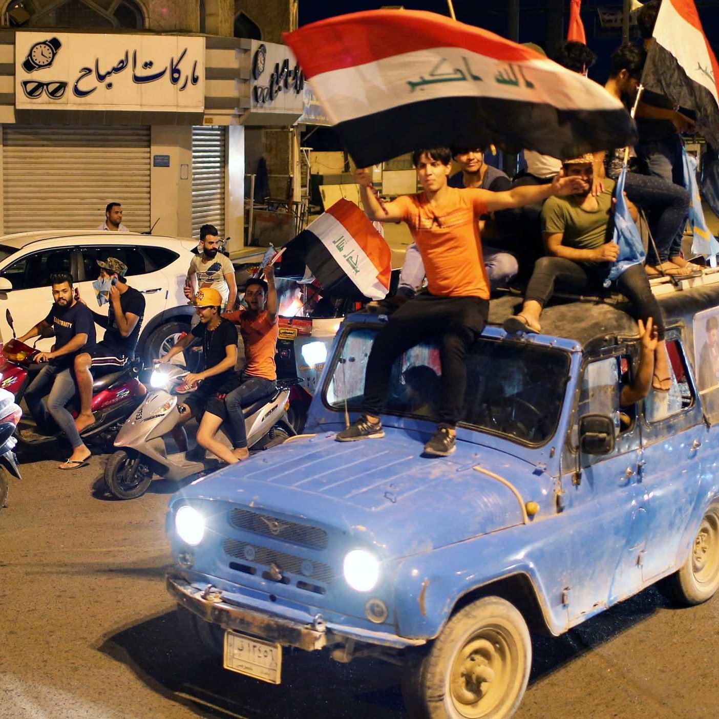 در این تصویر، گروهی از جوانان در یک خیابان در حال شادی و جشن هستند. آنها بر روی یک خودروِ آبی رنگ نشسته‌اند و پرچم عراق را در دست دارند. تعدادی موتورسیکلت نیز در کنار خودرو در حال حرکت هستند. جوانان با شور و هیجان به جشن و شادی پرداخته و فضای پرانرژی و شادابی را ایجاد کرده‌اند. در پس‌زمینه، ساختمان‌ها و فروشگاه‌هایی دیده می‌شوند که به شلوغی و نشاط این صحنه افزوده‌اند.