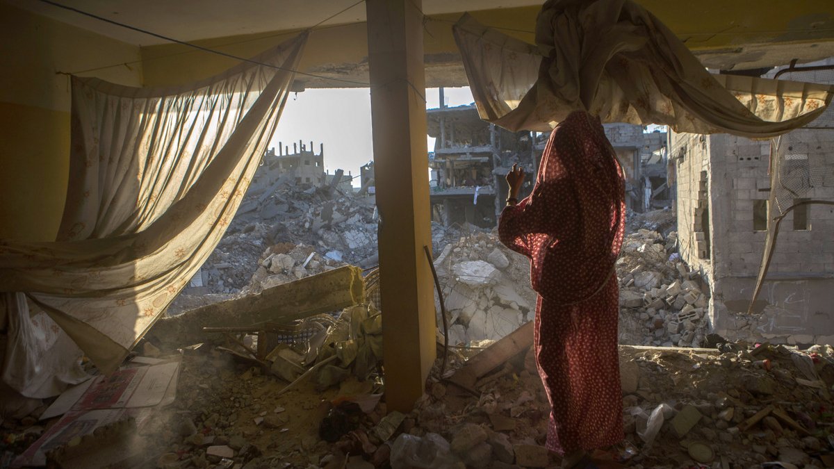 L'image montre une scène à l'intérieur d'un bâtiment endommagé. On aperçoit une femme vêtue d'un vêtement traditionnel, se tenant près d'une fenêtre qui donne sur des ruines. De l'autre côté, on voit des décombres de bâtiments effondrés, laissant entrevoir la désolation d'un environnement détruit. La lumière du soleil filtre à travers les rideaux, ajoutant une ambiance à la fois triste et poignante à la scène.
