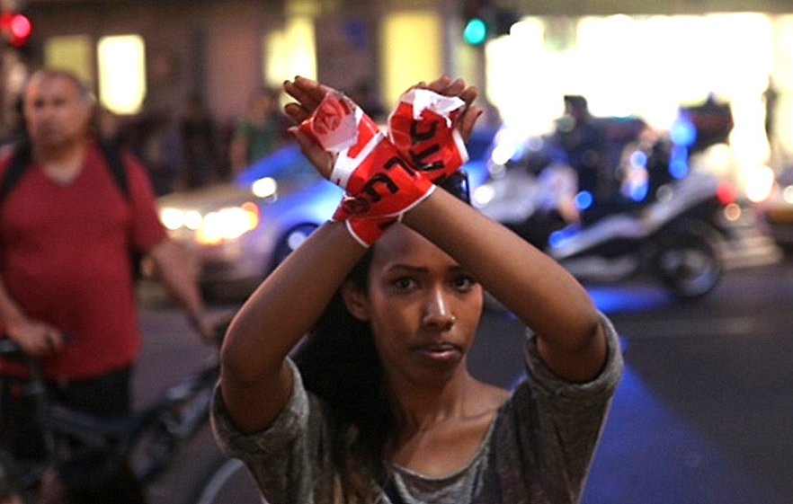 L'image montre une jeune femme qui se tient debout au milieu d'une rue, levant les bras avec des bandes de tissu rouge dans les mains, formant un symbole d'opposition ou de protestation. Elle semble déterminée et engagée, tandis que des véhicules et des lumières de police sont visibles en arrière-plan. Au fond, on peut apercevoir d'autres personnes, ajoutant un contexte urbain à la scène. L'atmosphère dégage un sentiment de mobilisation.