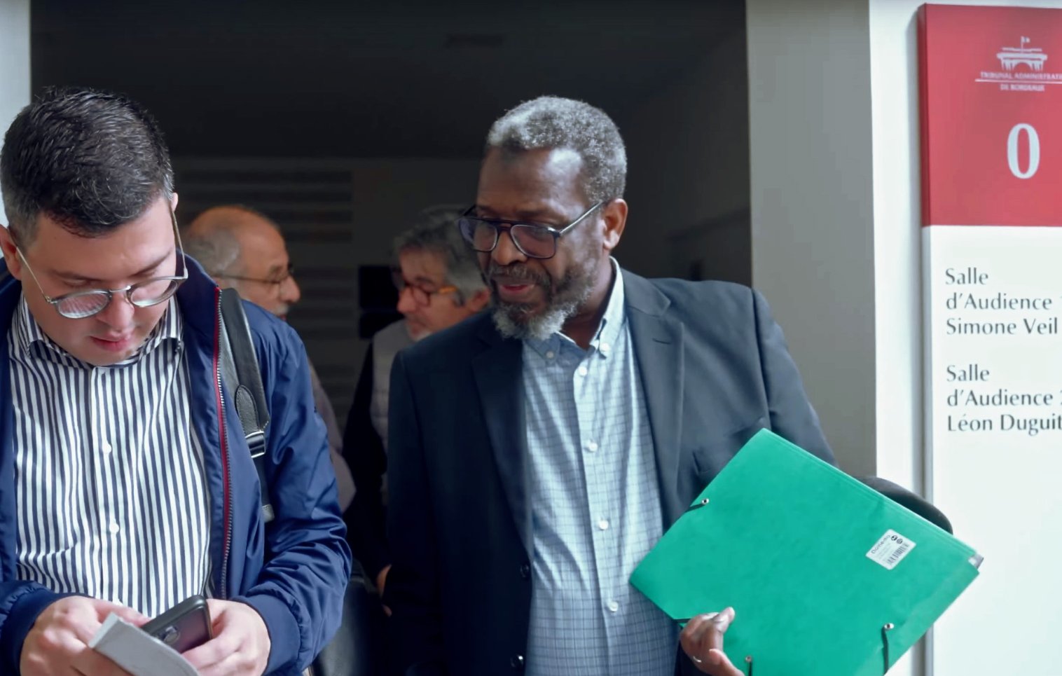 The image shows two men engaged in conversation as they exit a building. One man, wearing glasses and a navy jacket, is looking at his phone while the other, dressed in a black suit, is holding a green folder. In the background, there are other people visible, indicating a busy environment, possibly a court or conference setting. The signage indicates different audience rooms, adding context to the location.