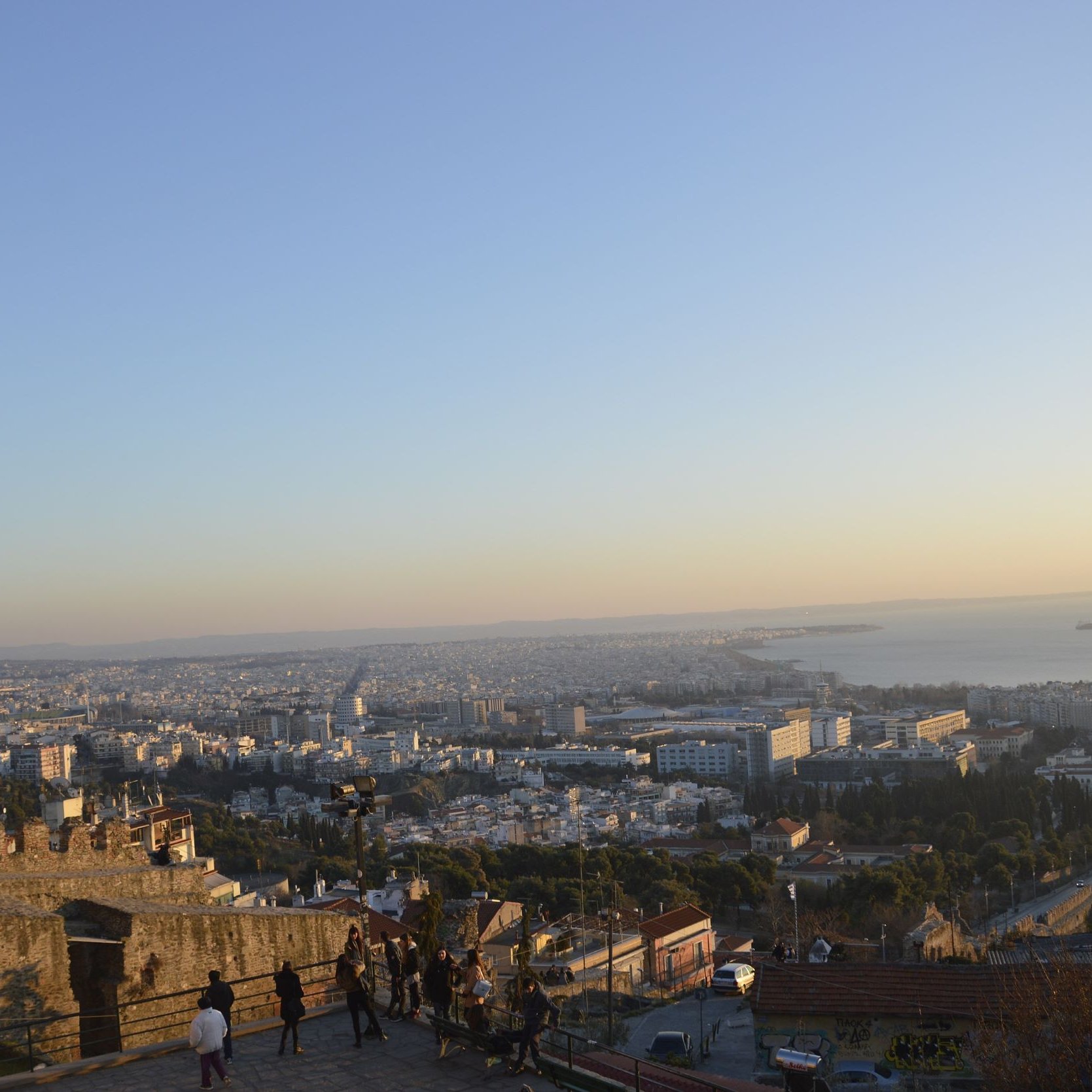 L'immagine mostra una vista panoramica di una città, probabilmente al tramonto. In primo piano si vede una parte di una muraglia o di una fortezza, con alcune persone che passeggiano. Sullo sfondo, si dispiega un vasto panorama urbano con edifici e strade, mentre il mare è visibile all'orizzonte, con alcune navi. I colori caldi del cielo creano un'atmosfera suggestiva, tipica delle ore serali.