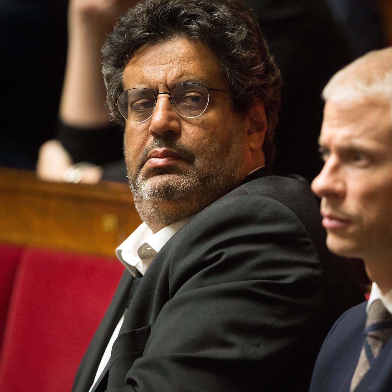 در این تصویر، چند نفر در یک جلسه یا مراسم رسمی نشسته‌اند. فردی که در جلوی تصویر قرار دارد، با موهای کوتاه و رنگ روشن، به دور نگاه می‌کند. در کنار او، فرد دیگری با موهای تیره و چهره‌ای جدی نشسته است. این صحنه فضایی جدی و رسمی دارد و افراد در حال توجه به موضوعات مطرح شده هستند. خلفیه تصویر کمی تار است و به نظر می‌رسد که دیگر افراد نیز در حال نشست هستند.