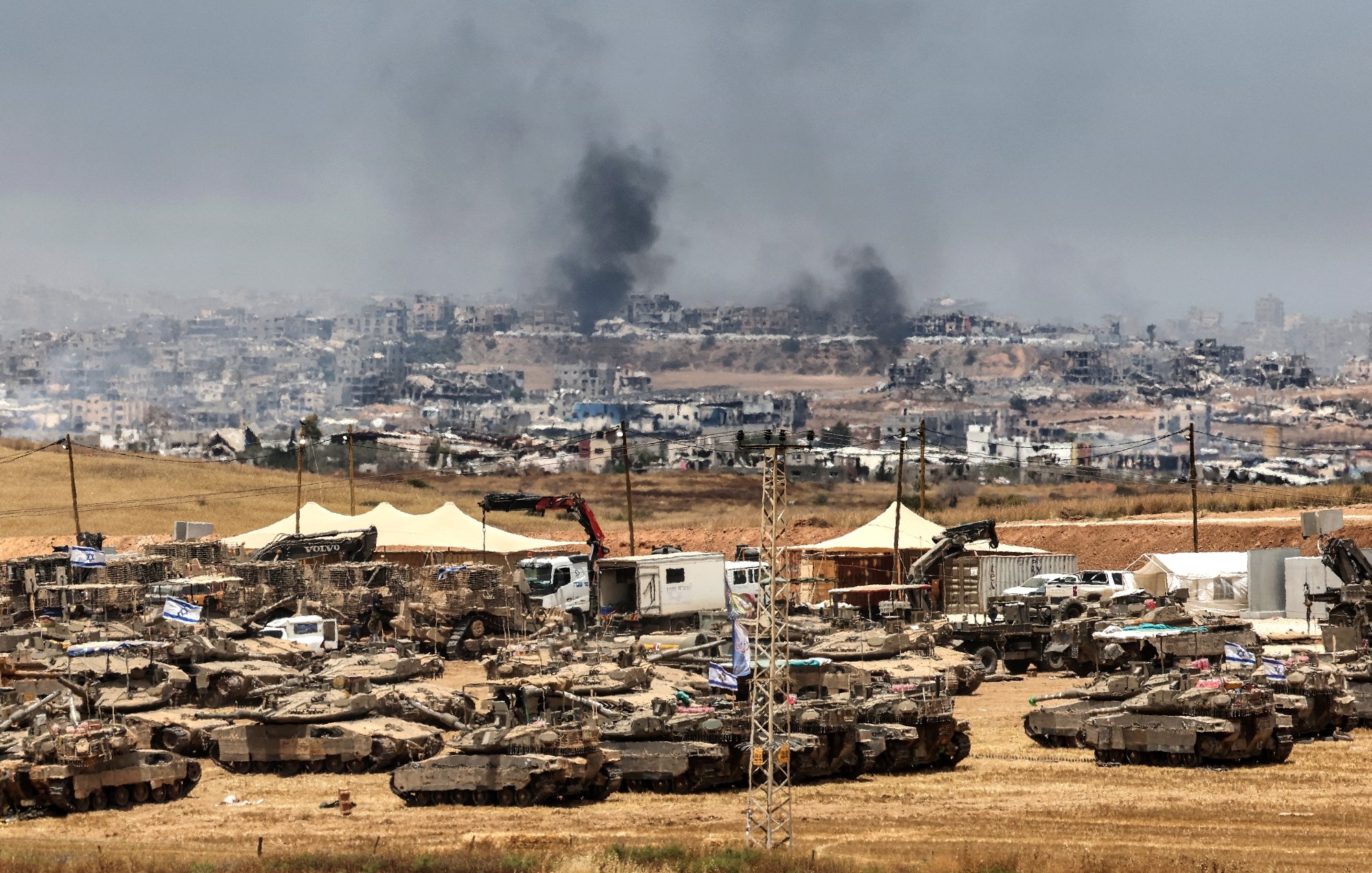 Un champ militaire avec des chars, fumée au loin et destructions visibles en arrière-plan.