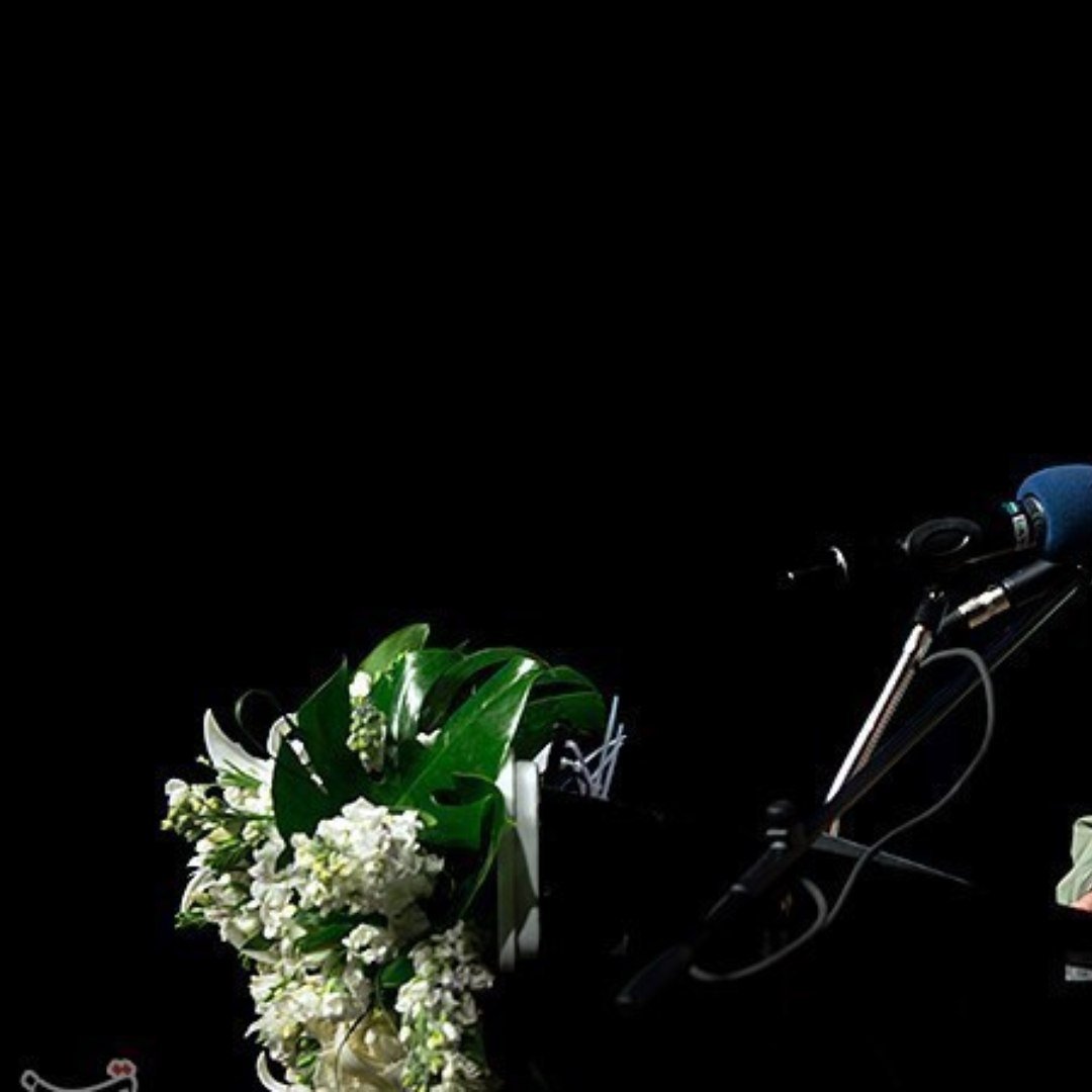 در این تصویر، یک فرد در حال سخنرانی در یک مکان رسمی است. او لباس نظامی به تن دارد و در حال بیان نکاتی با حرکات دست است. در کنار او، میزی با گل‌های تزئینی قرار دارد و میکروفون و بلندگو نیز دیده می‌شود. پس‌زمینه تصویر تاریک است و تمرکز بیشتری بر روی سخنران وجود دارد.