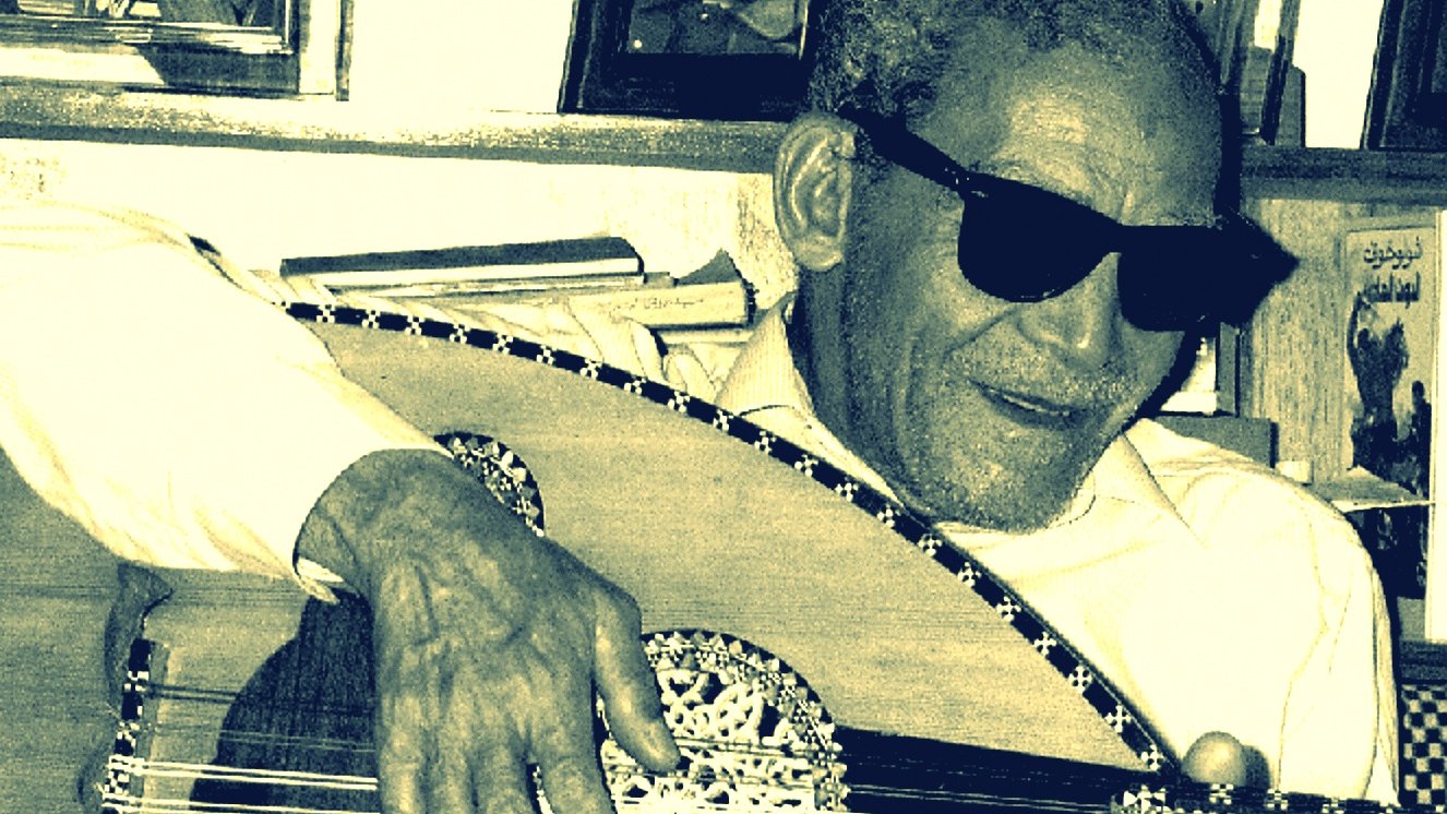 L'image montre un homme âgé portant des lunettes de soleil, assis et jouant de l'oud, un instrument de musique traditionnel. Il semble concentré sur sa musique, avec un sourire léger. En arrière-plan, on aperçoit des photos encadrées accrochées au mur, ce qui pourrait suggérer un environnement familial ou personnel. L'éclairage donne une ambiance nostalgique à la scène.