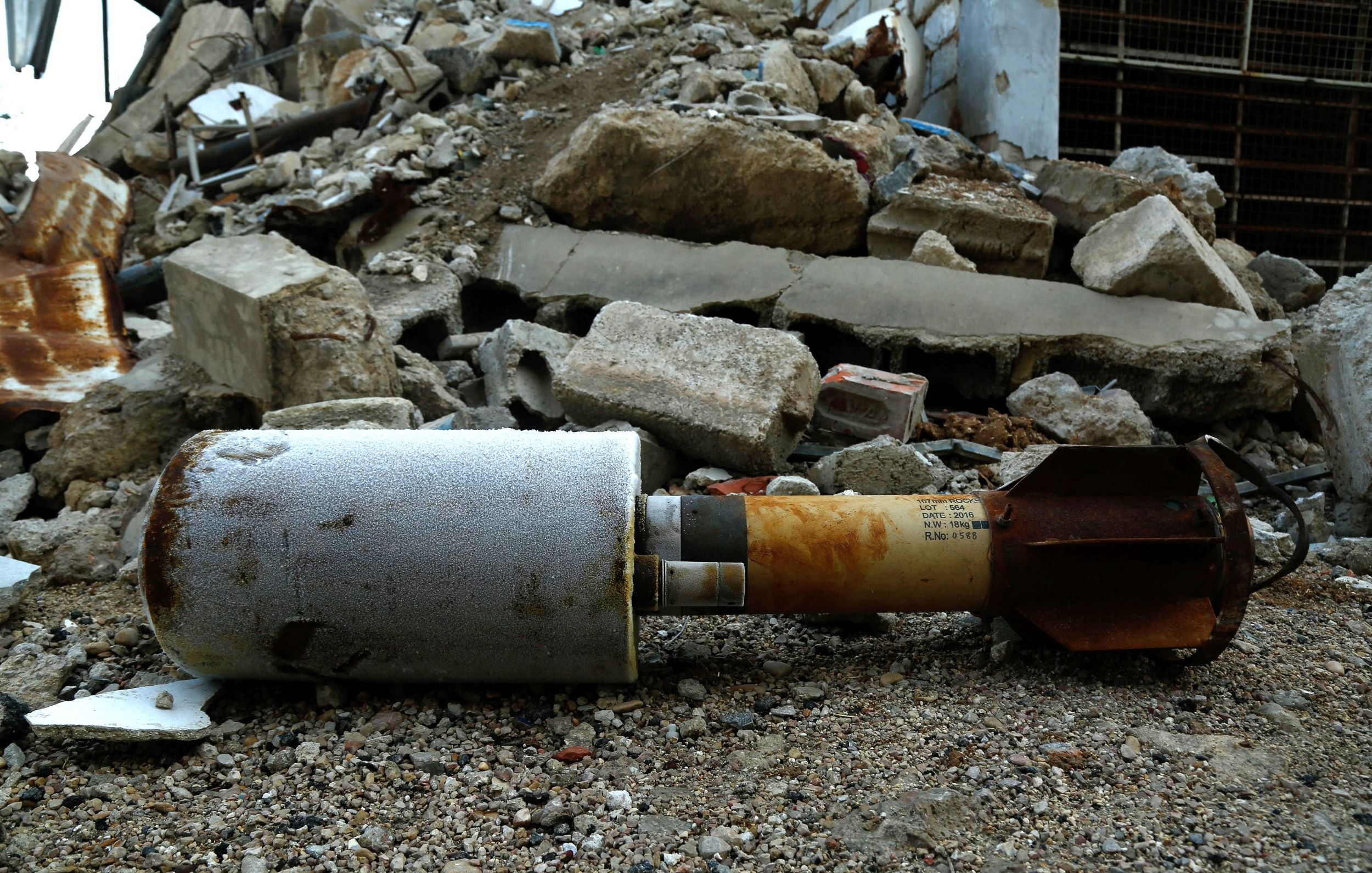 The image shows a missile or bomb lying on the ground among rubble and debris. The surrounding area is chaotic, with broken concrete and scattered materials suggesting destruction, possibly from an explosion or military action. The missile itself appears weathered and is partially covered in grime, indicating it has been in this location for some time.