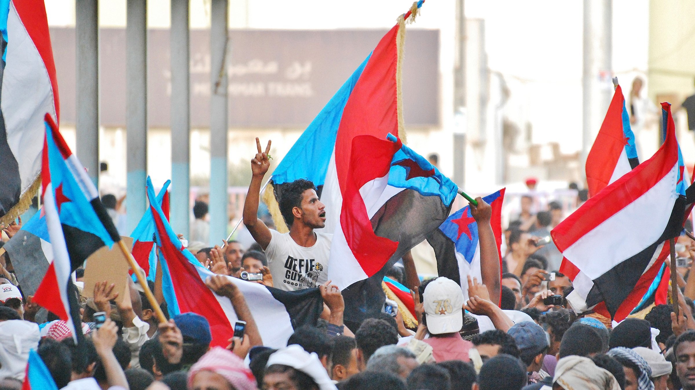 L'image montre une foule rassemblée, brandissant des drapeaux d'un pays ou d'une région, probablement en lien avec une démonstration ou une manifestation. Au centre, un homme se démarque en levant deux doigts en signe de victoire. La scène est animée, avec beaucoup de personnes exprimant leur enthousiasme ou leur engagement, ce qui indique une ambiance de protestation ou de célébration. Les couleurs des drapeaux sont vives, ce qui ajoute à l'énergie de la scène.