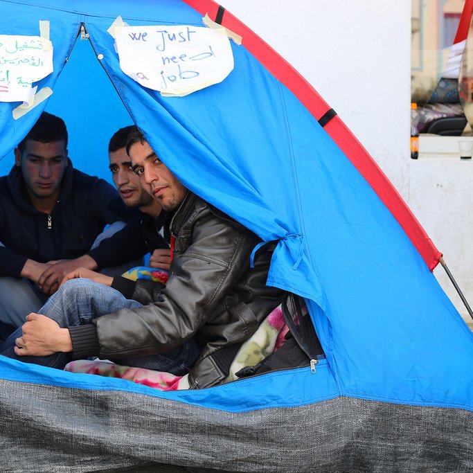 L'image montre un groupe de trois jeunes hommes assis à l'intérieur d'une tente. Ils semblent discuter ensemble, et des pancartes en arabe et en anglais sont visibles à l'extérieur de la tente, indiquant "Nous avons juste besoin d'un emploi". La tente est de couleur bleue, et l'ambiance générale évoque un sentiment de rassemblement autour d'une cause.