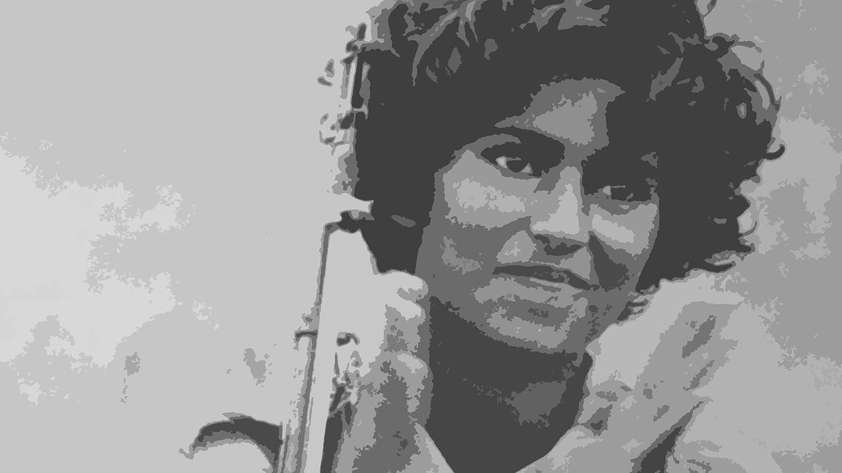 L'image montre une femme avec des cheveux bouclés, tenant un fusil. Elle sourit légèrement tout en applaudissant, ce qui semble indiquer un moment de célébration ou de camaraderie. Son uniforme militaire est visible, ce qui suggère qu'elle est impliquée dans un contexte de lutte ou de conflit. L'arrière-plan est flou, mettant l'accent sur sa figure.