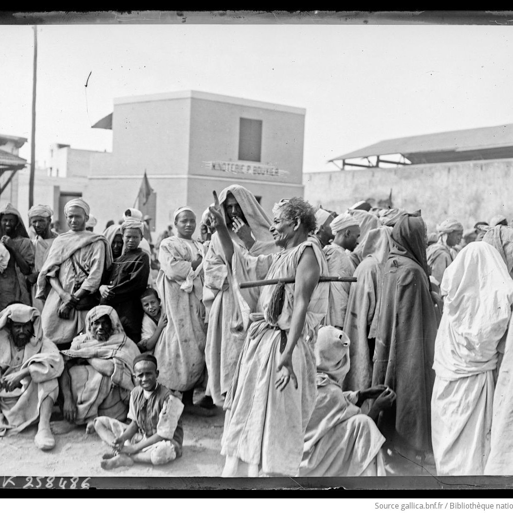 La imagen muestra a un grupo de personas en un entorno que parece ser un mercado o una reunión al aire libre. La mayoría de las personas están vestidas con ropas tradicionales, muchas de las cuales son de colores claros y están cubiertas con prendas que podrían indicar el clima cálido de la región. Hay un enfoque particular en una figura central que parece estar gesticulando o hablando, rodeada de un público atento. El ambiente es de interacción social y hay una variedad de expresiones en los rostros de las personas, que van desde la curiosidad hasta la concentración. El fondo muestra edificaciones simples, lo que sugiere un contexto urbano o rural.