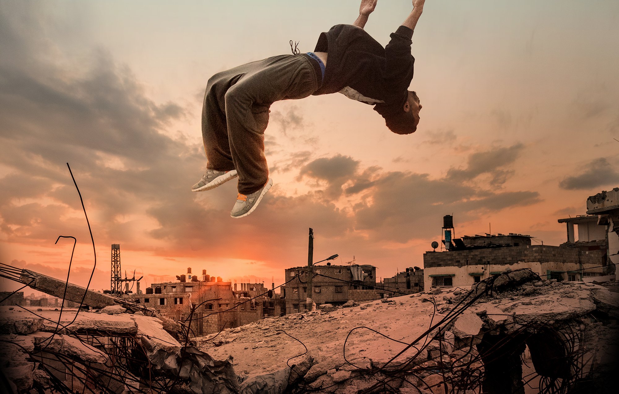 L'image montre un jeune homme en train de sauter dans les airs, effectuant une figure acrobatique, au-dessus des ruines d'une ville. En arrière-plan, le ciel est coloré de teintes orangées et bleutées, ce qui donne une ambiance dramatique à la scène. Les débris et les structures en ruine témoignent d'un environnement urbain dégradé, ajoutant un contraste frappant avec la dynamique du saut.